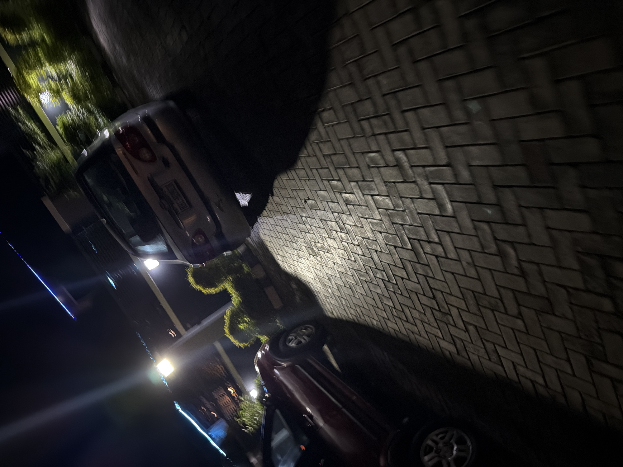 The image shows a dark paved driveway at night, with two cars parked on it. A silver car is parked on the right, facing away from the camera. A red car is parked on the left, facing towards the camera. The red car appears to be a hatchback, and the silver car is a sedan. The driveway is made of interlocking brick pavers, and there are some bushes and trees in the background. A light source, possibly a streetlamp, is casting a strong shadow from the red car onto the driveway. The scene appears to be in a residential area.