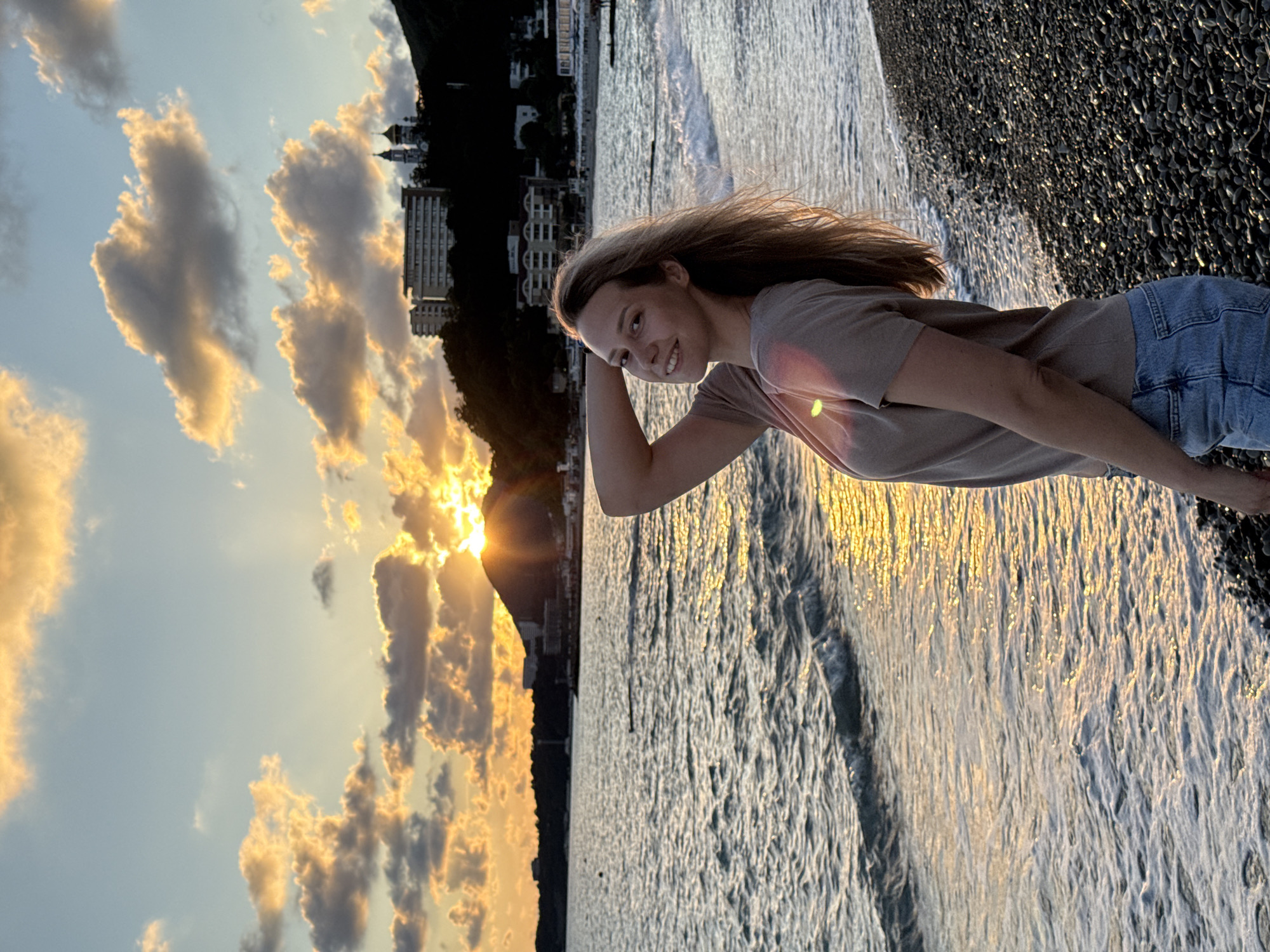 A woman with long blonde hair stands on a pebble beach in Nebug, Russia, at sunset. She is wearing a tan t-shirt and blue denim shorts. Her right arm is bent behind her head, and she is looking at the camera with a smile. The sea is in front of her, with gentle waves lapping at the shore. The sun is setting behind a mountain in the distance, casting a warm glow over the scene. Buildings and a church with golden domes are visible on the hillside. The sky is filled with dramatic clouds, tinged with orange and yellow from the setting sun.