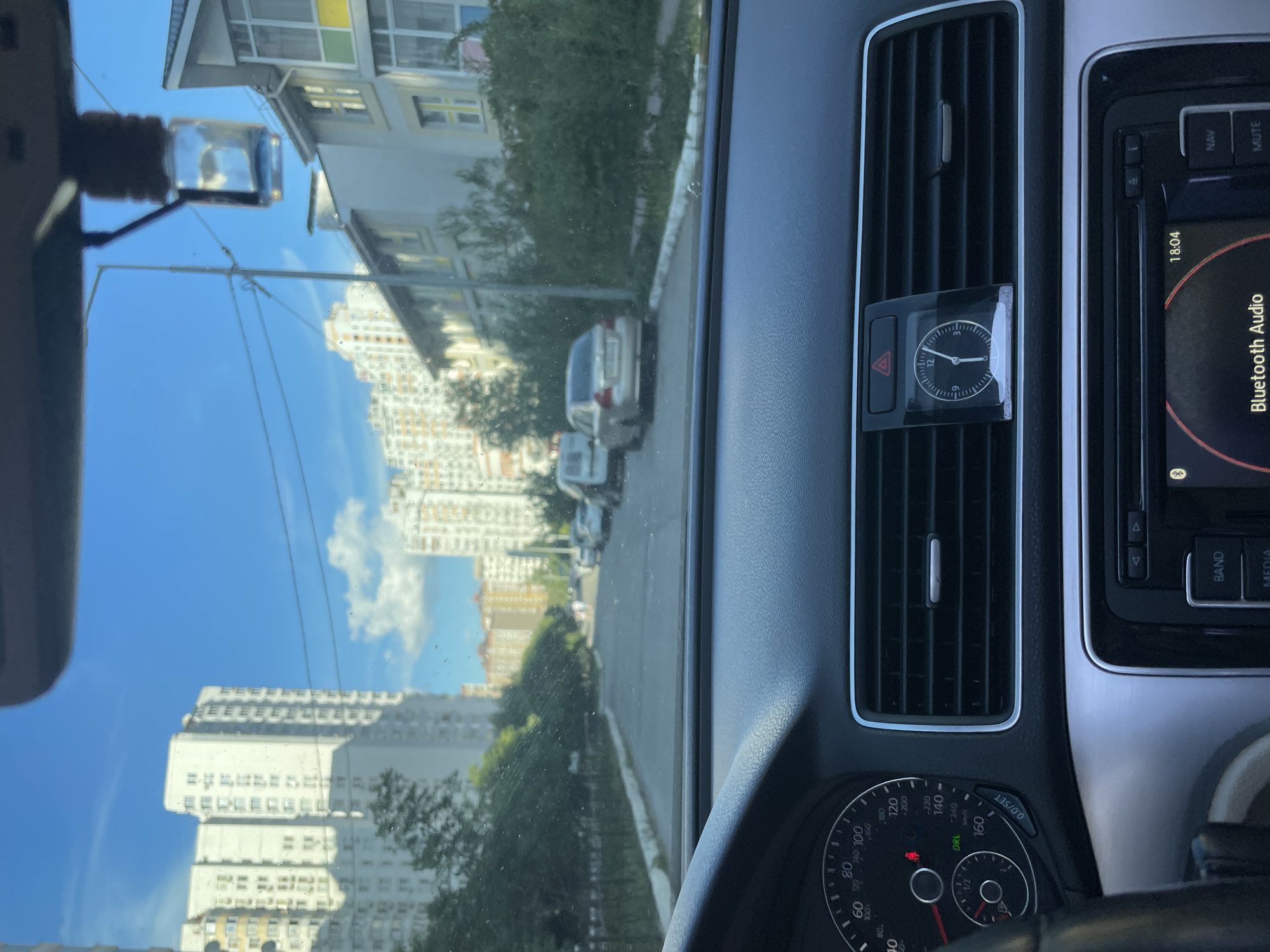 The image, taken from inside a car in Kyiv, Ukraine, shows a street scene dominated by modern apartment buildings under a bright blue sky with scattered clouds. The car's dashboard is partially visible, with a speedometer on the left and a dashboard clock, showing approximately 12:05, placed above an air vent. Several cars are parked or driving along the asphalt road. The overall impression is one of a residential area in a contemporary urban environment.