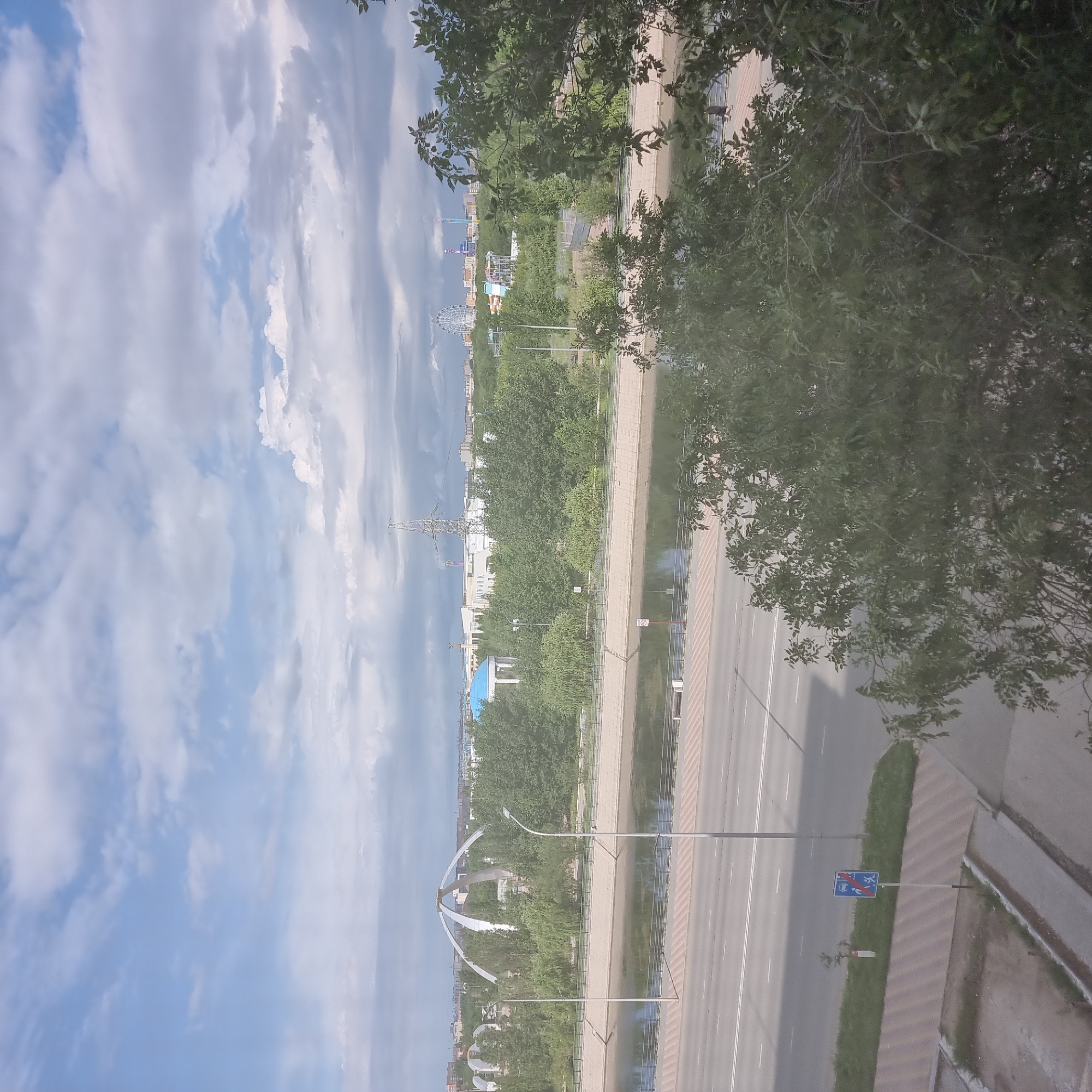The image captures a panoramic view of a cityscape, likely from an elevated vantage point, showcasing a river, a road, and various structures against a cloudy sky.
In the foreground, a dense canopy of green foliage partially obscures the view of the scene below. A paved road with distinct white lane markings runs horizontally across the lower portion of the image, with a grassy area and a sidewalk adjacent to it. A blue and red traffic sign with symbols indicating vehicles and pedestrians is visible on the side of the road.
Further back, a calm river flows parallel to the road, its surface reflecting the sky and surrounding environment. The riverbanks are lined with concrete embankments and a pathway. Beyond the river, a treeline dominates the mid-ground, with various types of trees creating a green buffer.
In the distance, several architectural landmarks are discernible. A prominent white, curved structure resembling an arch or a large gateway stands out amongst the trees on the left side of the frame. Further into the city, a tall, lattice-like transmission tower is visible, alongside buildings with blue and white roofs. To the right, a Ferris wheel and other amusement park-style structures are present, suggesting a recreational area within the city. The overall impression is of an urban landscape interspersed with natural elements and man-made attractions.
The sky is filled with a mix of light and dark clouds, with patches of blue sky peeking through, indicating variable weather conditions. The lighting suggests it is daytime, likely late morning or afternoon.
The provided context indicates the location is the city of Aktobe, Kazakhstan. This scene appears to be a view of the embankment of the Ilek River, a significant geographical feature in Aktobe. The large white arch is likely a monument or part of a park along the river. The transmission tower and amusement park rides are common urban features that would be found in a city of Aktobe's size.