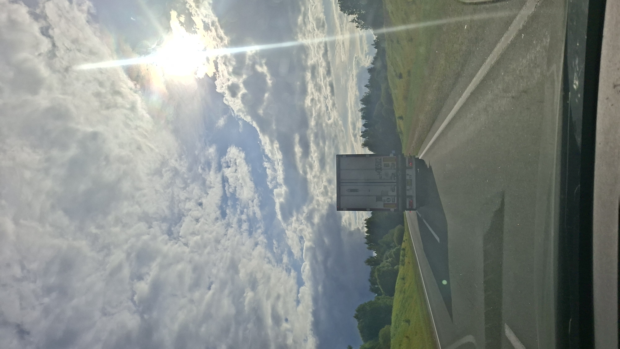 The image captures a view from a vehicle traveling on a highway in Верхнякрывінскі сельскі Савет, Belarus. The road ahead is paved and has white lane markings. A white semi-trailer truck is visible in the distance, traveling in the same direction. The truck has markings on its rear, including the text "H00502" and other symbols.
The landscape on either side of the highway consists of grassy hills with scattered trees. The sky is filled with a mix of clouds, with a bright sun visible towards the upper right, casting a strong beam of light. The lighting suggests it is daytime, likely late afternoon or early evening due to the angle of the sun. The overall scene depicts a rural highway environment under a cloudy sky.