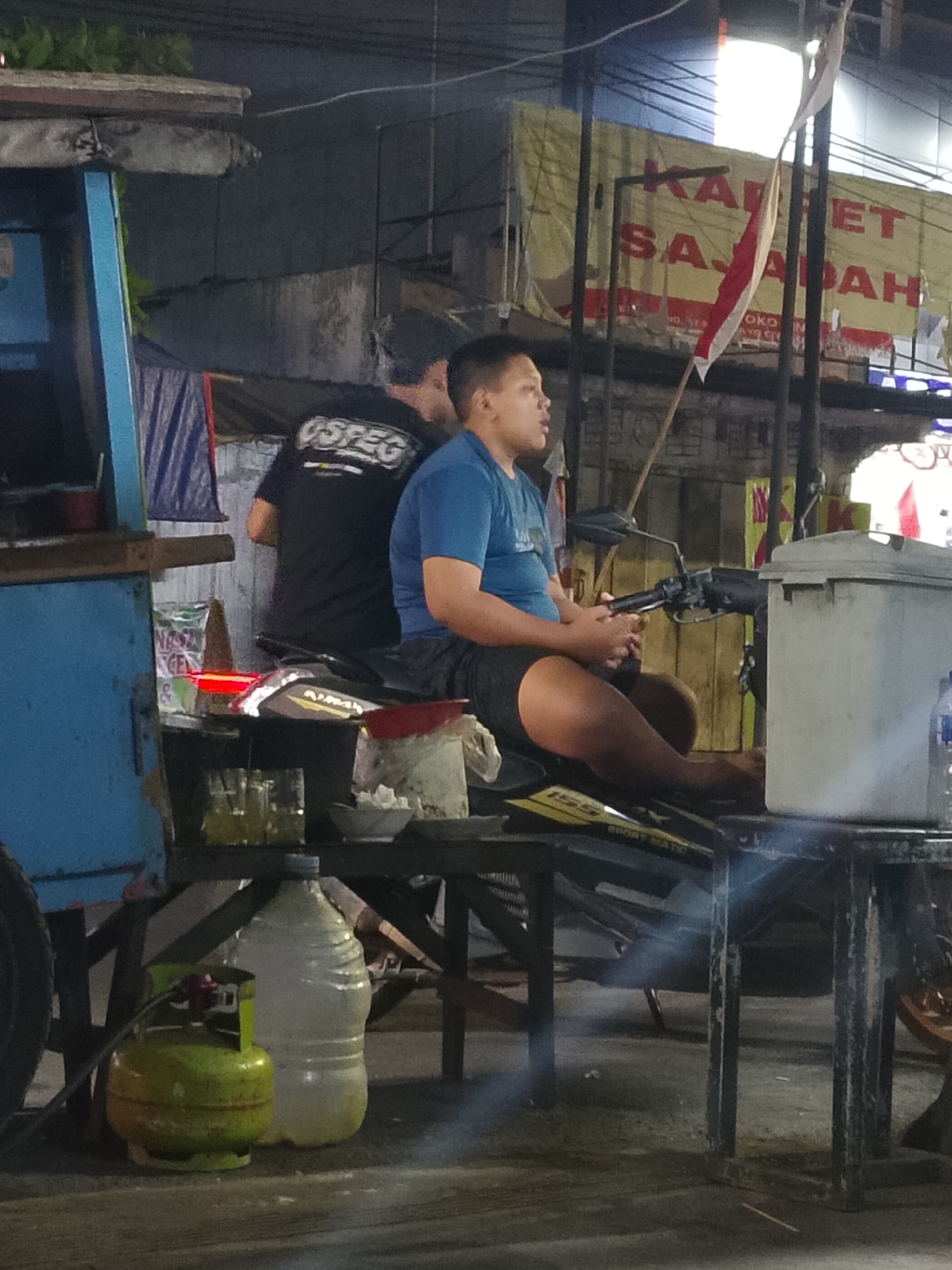 In Balun, Indonesia, at night, a young male wearing a blue t-shirt and dark shorts sits on a motorcycle. To his left, another male is partially visible, wearing a black t-shirt with "OSPEC" printed on the back. The background features a yellow banner with red text that reads "KARPET SAJADAH". A red and white Indonesian flag hangs near the banner. In the foreground, a blue food cart is on the left. On a wooden table in front of the motorcycle are several glasses with liquid, a bowl with white cubes, and a large plastic bottle filled with water. A green gas cylinder is also visible on the ground near the food cart. The lighting suggests an outdoor setting at dusk or night, possibly near a street or market.
