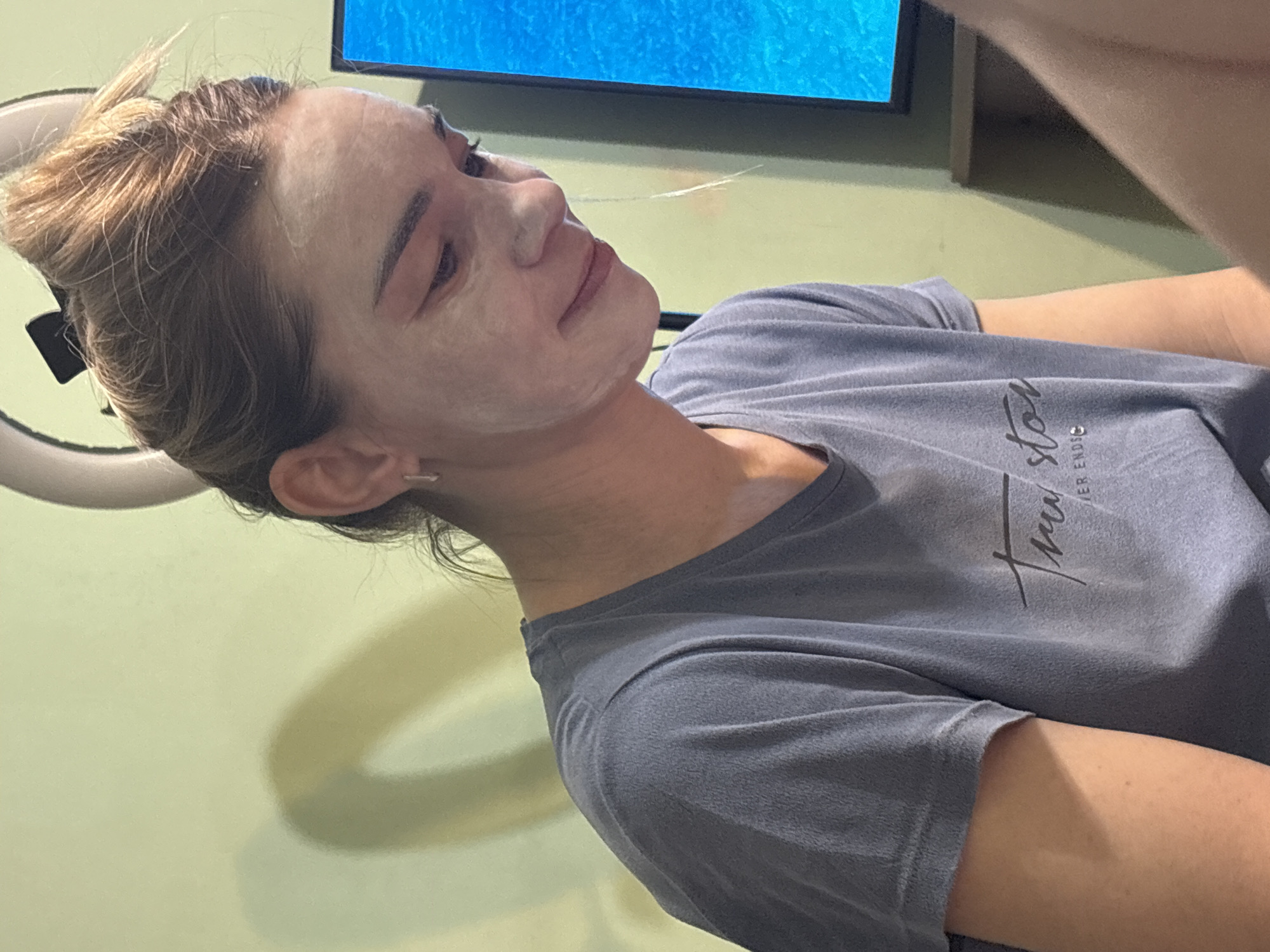 A woman with light brown hair pulled back in a bun is visible from the chest up. Her face is covered in a white substance, possibly a mask. She is wearing a gray t-shirt with lettering on the front. Behind her is a green wall and a television screen displaying blue imagery. A ring light is also visible to the left of her head. The location of the image is not indicated.