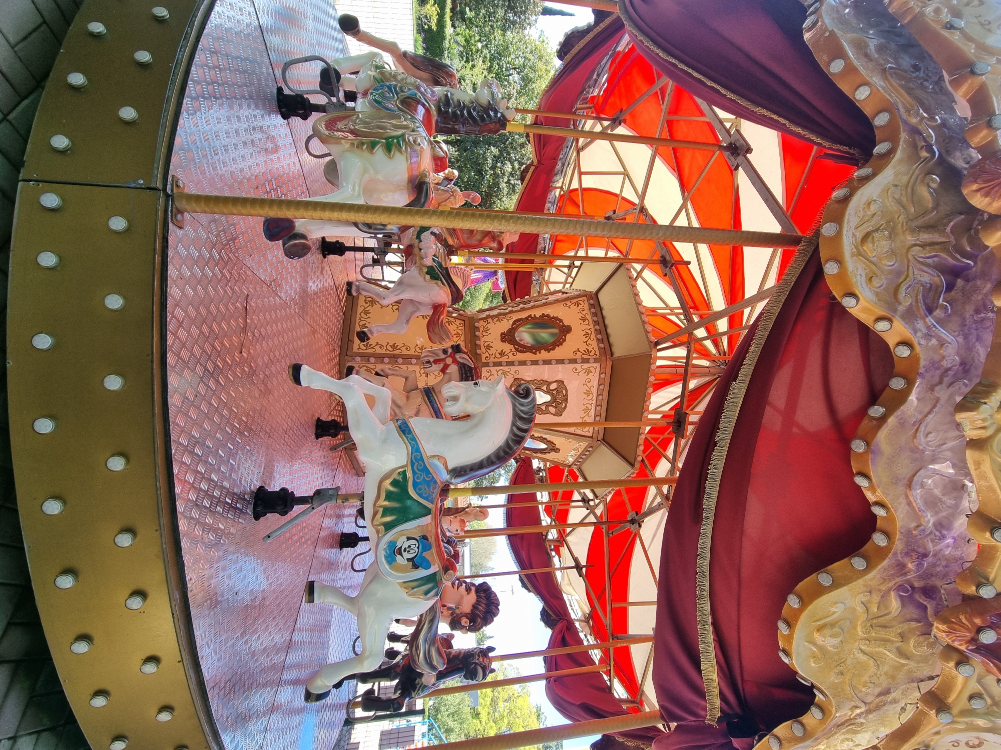 The image depicts a carousel in Sochi, Russia. The carousel is adorned with red and white striped awning panels, partially draped with maroon curtains trimmed with gold fringe. The carousel features painted horses, some white with blue and green accents, others in brown tones. There are also seats or carriages with ornate designs. The floor of the carousel has a patterned, possibly metallic, surface. The outer edge of the carousel is decorated with lights. The background shows green trees and a glimpse of buildings, typical of an urban park setting.