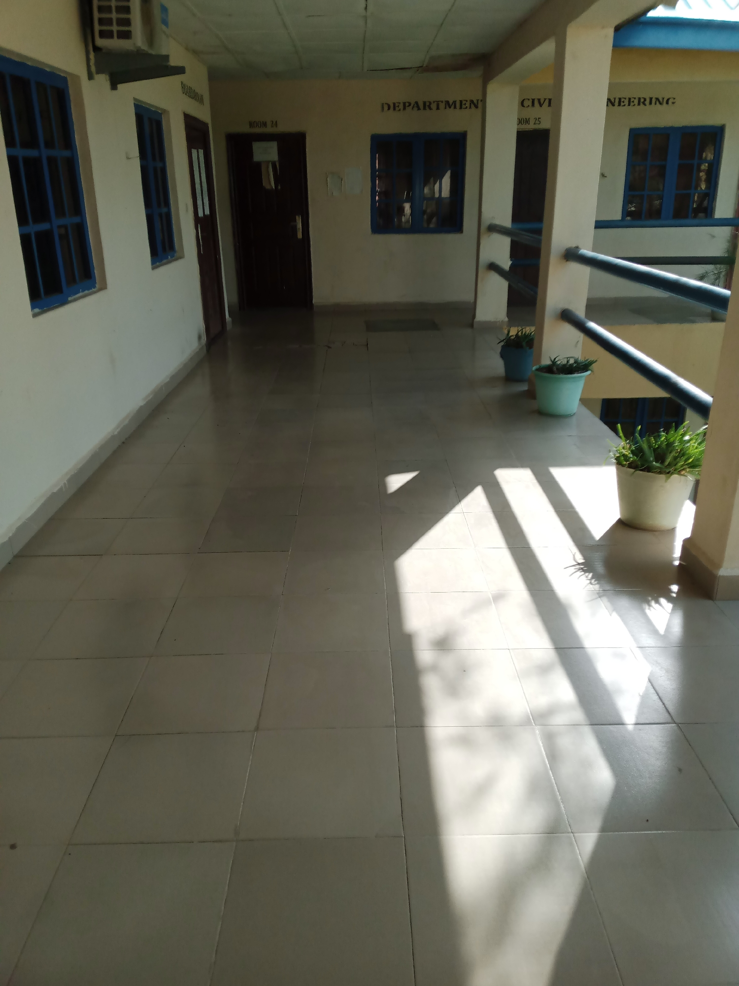 An outdoor walkway with a tiled floor shows a sunlit corridor with the shadows of a railing cast across the floor. The walkway is flanked by a cream-colored wall on the left, featuring blue-framed windows. On the right, the walkway opens to a view of the building's interior. Signs on the wall indicate "BOARDROOM" and "ROOM 24" on the left, and "DEPARTMENT", "CIVIL ENGINEERING", and "ROOM 25" on the right. Several potted plants are placed along the railing on the right side of the walkway. The overall impression is one of a functional, institutional building, likely part of a school or office complex in Jalingo, Nigeria.