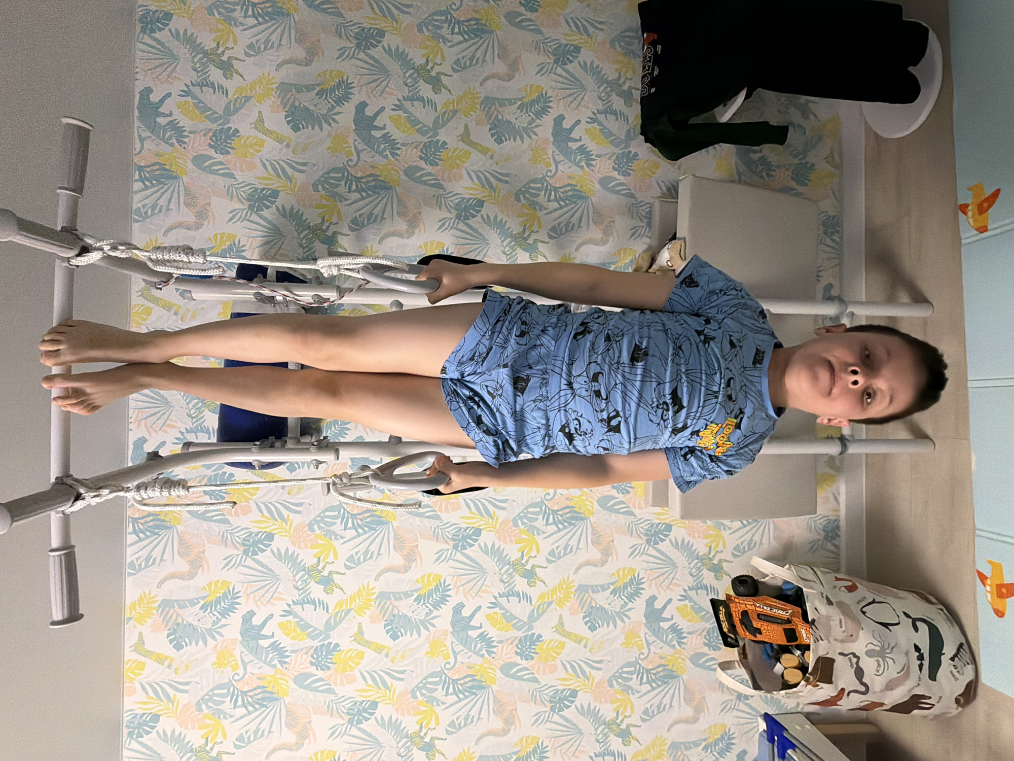 A child in a blue patterned pajama set lies on a traction bed in a room. The room is decorated with a wallpaper pattern of dinosaurs and jungle leaves. Beside the bed there is a bag filled with toys. This image was taken in Votkinsk, Russia.