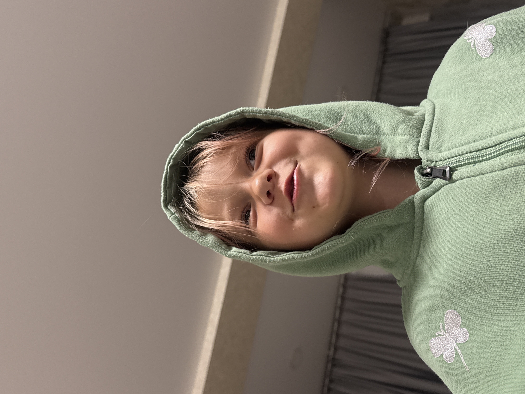 A young girl with blonde hair and a light green hooded jacket is pictured from a low angle looking up. The girl has a slight smile and her eyes are looking upwards and to her right. She has a smudge of pink or red on her upper lip. The background is out of focus, showing a ceiling with a light fixture and a grey curtain in the distance. The jacket has a zipper closure and a silver butterfly design on the left breast. The location is not specified.