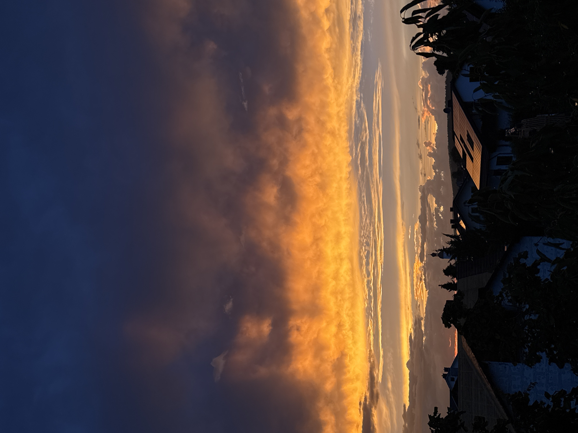 The image captures a dramatic sunset over the town of Wettstetten, Germany. The sky is filled with a vibrant display of orange, yellow, and dark blue clouds, creating a stark contrast. In the foreground, the silhouettes of rooftops and trees are visible, obscuring some of the details of the town. A church spire can be seen rising above the rooftops, indicating the presence of religious buildings within the community. The overall scene evokes a sense of twilight and natural beauty, with the setting sun casting a warm glow over the landscape.