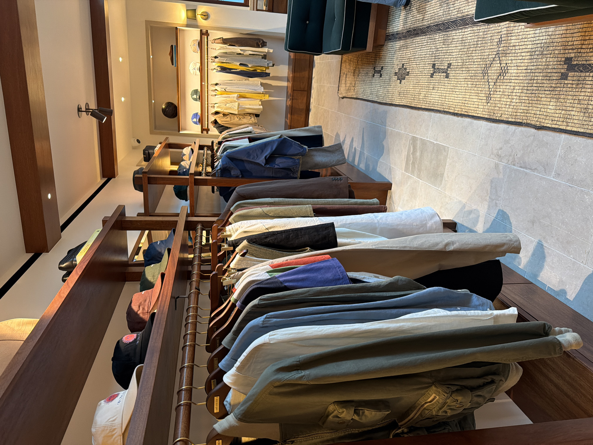 The image captures the interior of a retail space, seemingly a clothing store, with racks of apparel neatly displayed on wooden shelves and hangers. Predominantly, the focus is on the clothing, with a variety of colors and fabrics suggesting a range of styles. The store's interior design incorporates wooden fixtures and a neutral color palette, creating a warm and inviting atmosphere. In the background, there's a mirror reflecting additional clothing displays and a glimpse of seating. The flooring appears to be a light-colored tile, and a patterned rug adds texture and visual interest to the space. Given the upscale and curated aesthetic of the store, it's plausible to infer that the location could be in West Hollywood, USA, which is known for its boutique shops and design-forward retail environments.
