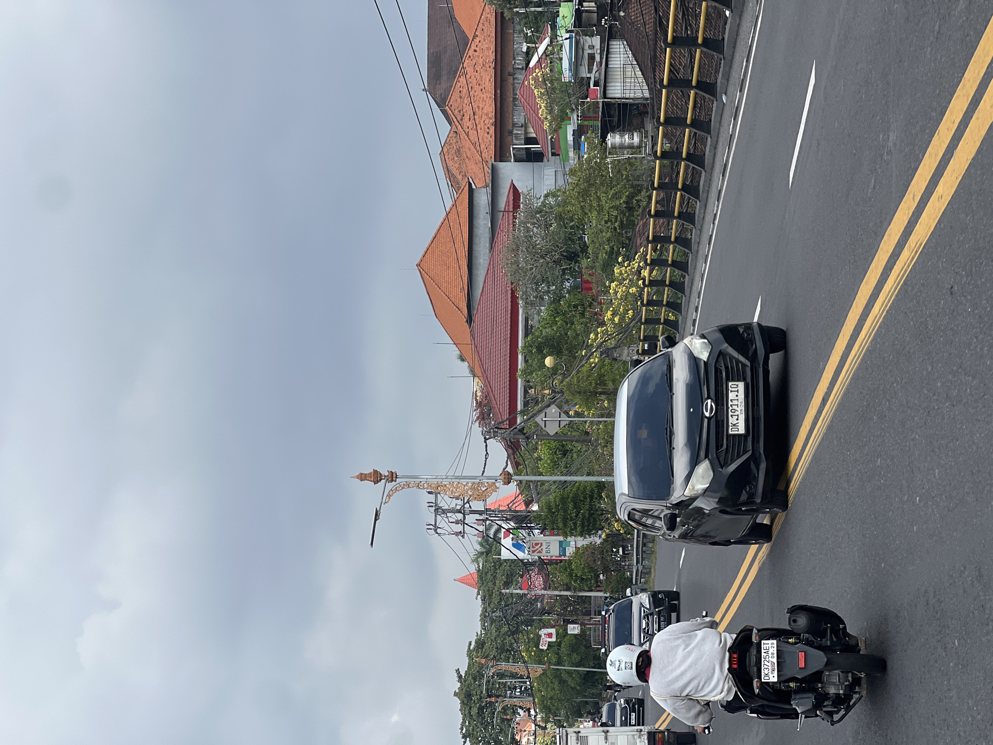 This is a vertical, eye-level, medium shot of a traffic scene on a cloudy day in Denpasar, Indonesia. In the foreground, a person wearing a white helmet and a light-colored long-sleeved shirt rides a black scooter down a wet, asphalt road marked with yellow double lines. The scooter has a license plate that reads "DK 3725 AET". To the right of the scooter, a black car with a license plate "DK 1911 IQ" is in the adjacent lane. Further down the road, other vehicles are visible. Along the right side of the road, a bridge railing with yellow posts and black railings is present, beyond which are buildings with red-tiled roofs and lush green trees. Streetlights with ornate, gold-colored decorations are seen above power lines. In the background, more buildings with similar roofs and a cloudy sky complete the scene.