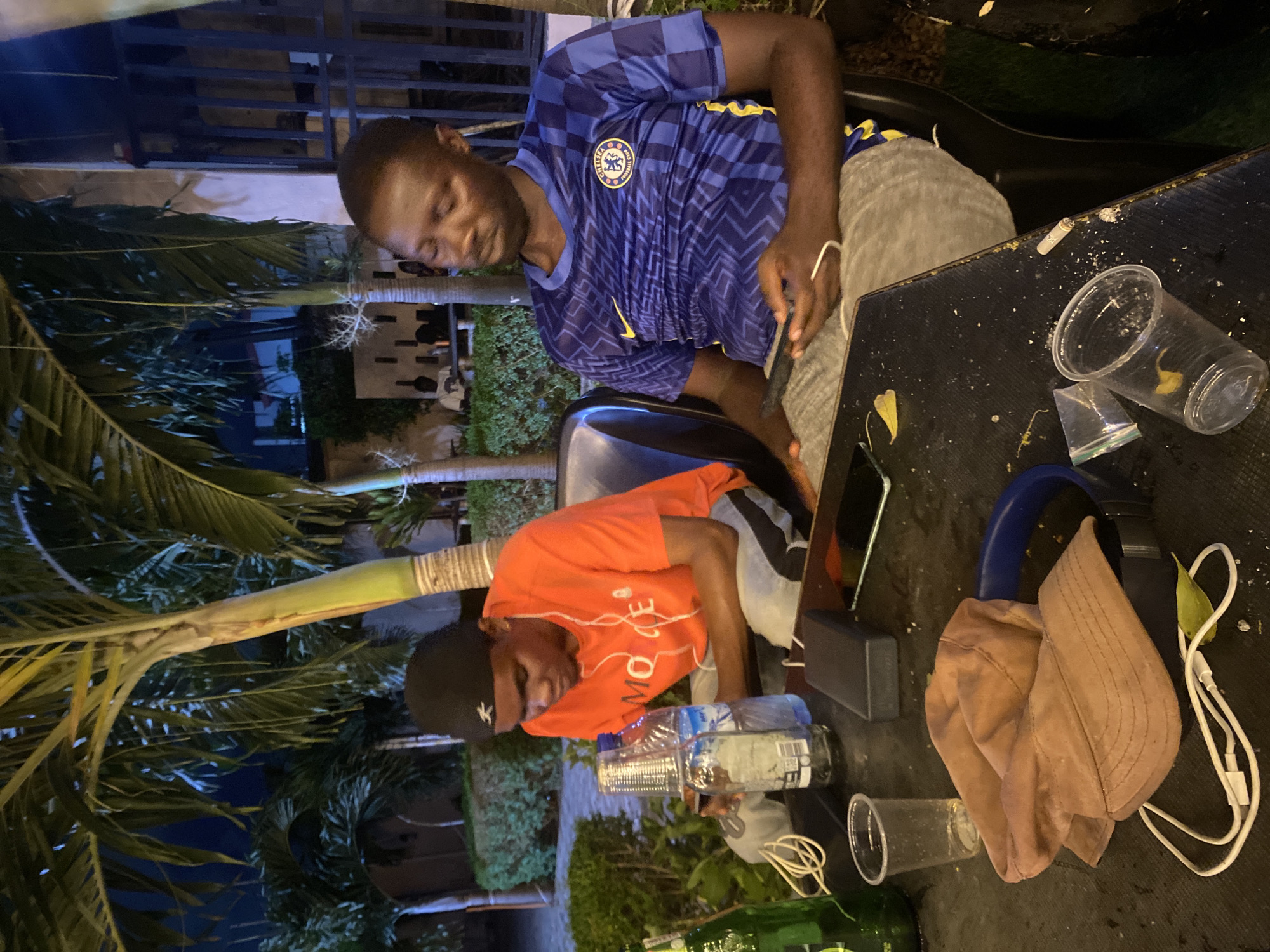 The image captures an outdoor evening scene. Two men sit near a black table littered with items. One man wears a blue Chelsea football jersey and grey pants, appearing tired, with his head slightly tilted. The other man wears an orange shirt and grey pants with a black cap. On the table are several plastic cups, water bottles, a smartphone, a portable charger, a pair of headphones, and a tan-colored hat. A cigarette butt rests on the edge of the table. Palm trees and city lights are visible in the background, suggesting a location in Lagos, Nigeria.
