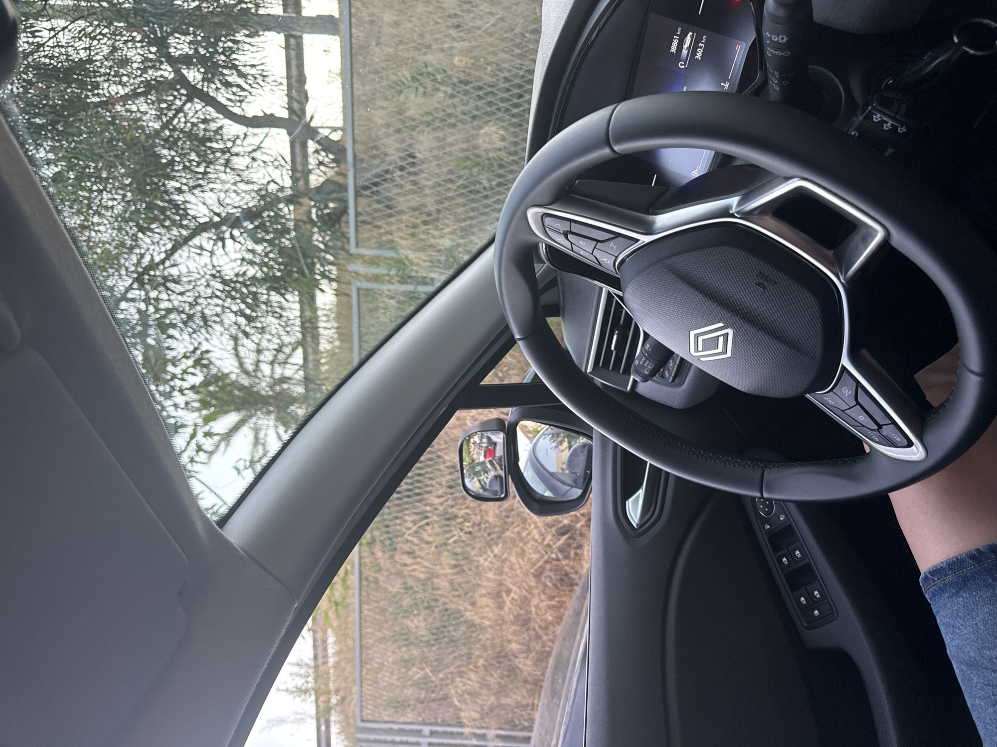 The image shows the interior of a car, looking out the windshield and side window. A person's legs, wearing denim shorts, are visible on the left side of the frame. The steering wheel, with a logo in the center and controls on the sides, is prominent. The car's dashboard shows a digital display with "38861 km" and "360.3 km". Outside the car, through the window, a metal fence and dry vegetation are visible, with trees and a bright sky beyond. A side mirror is also visible, reflecting a red car. The scene appears to be in a parking area or along a road with an outdoor environment. The image does not provide any specific information to determine the location in the city of Murcia, Spain.