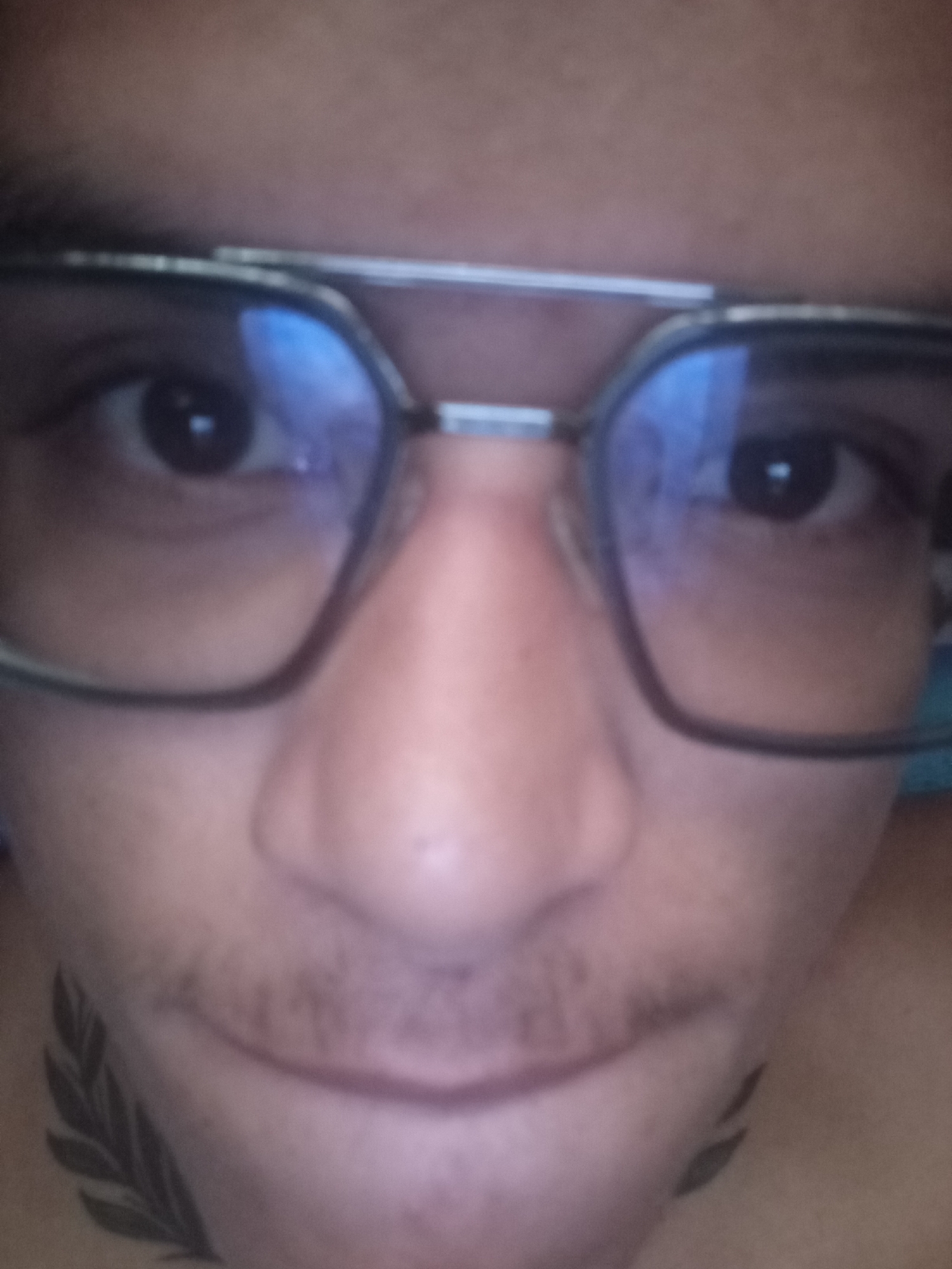 A close-up image of a person's face. The person is wearing glasses with a silver frame. Their eyes are dark and visible through the lenses. The bridge of their nose is prominent. A thin mustache is present above their upper lip. A tattoo resembling a leafy branch is visible on the left side of their neck, near the jawline. The background is out of focus. Location and further context are unavailable.