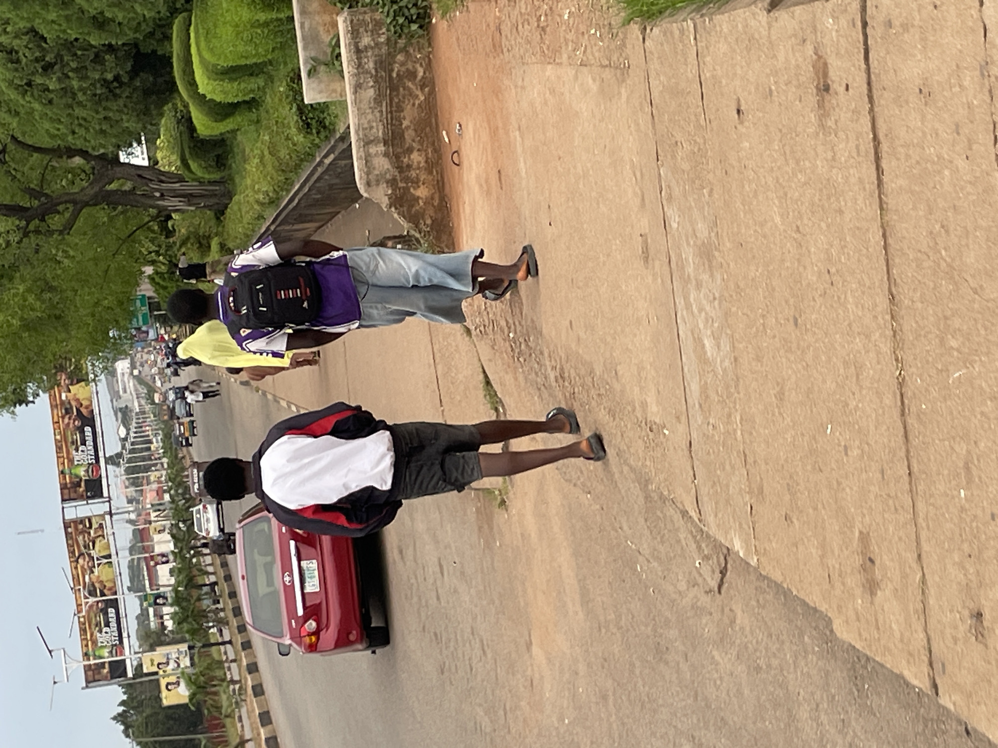 This image captures a scene on a roadside in Jos, Nigeria. Two young men are walking away from the camera on a paved sidewalk. The man in the foreground is wearing a white and red jacket over dark shorts and flip-flops. The man behind him wears a purple and white shirt, light blue denim shorts, a black backpack, and flip-flops. They are walking along a sidewalk bordering a busy road with traffic, including a red car and other vehicles visible in the background. A large billboard with advertisements is mounted on poles over the road. The environment includes lush green trees and meticulously trimmed hedges lining the sidewalk, suggesting a well-maintained urban area. The sky is overcast.