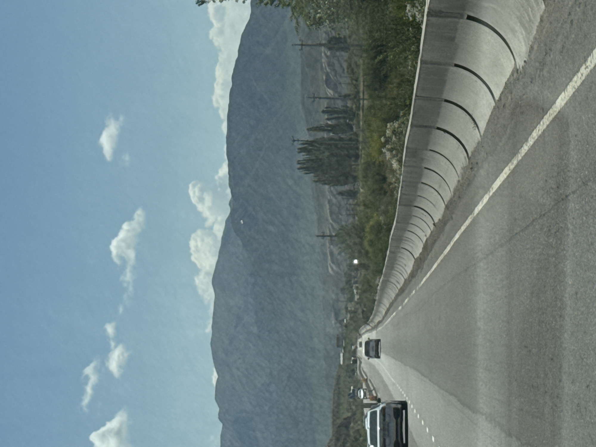 The image depicts a highway in or near Krasnyy Most, Kyrgyzstan. The foreground shows a paved road with lane markings, and a concrete barrier on the right side. Several vehicles are visible on the road, including a white SUV on the left and a darker car in the distance. The middle ground features sparse vegetation and several tall, slender trees. In the background, a large, blue-gray mountain range dominates the horizon under a bright blue sky with scattered white clouds. Power poles are also visible along the road.