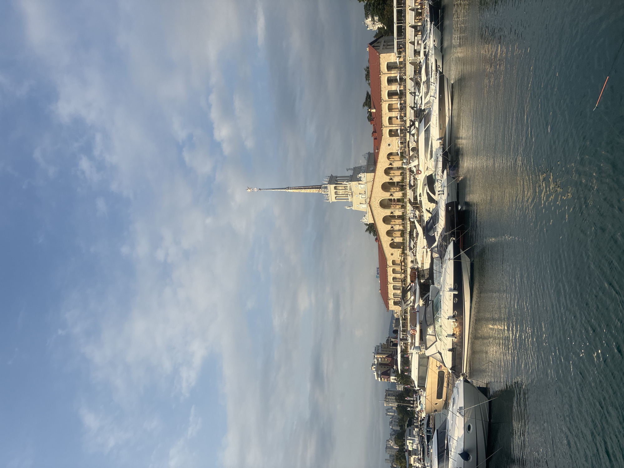The image shows the Sochi Sea Port in Sochi, Russia. It is a sunny day with scattered clouds in a blue sky. The port is filled with numerous yachts of various sizes, docked along the quay. In the background, a large, ornate building with a tall, spire-like tower stands prominently. The building has a long, arched facade and a red-tiled roof. The water in the harbor is calm and reflects the surrounding structures and the sky. Buildings and trees can be seen in the distance, suggesting a city setting. There are no people clearly visible in the image. The overall scene depicts a vibrant and well-maintained maritime hub.