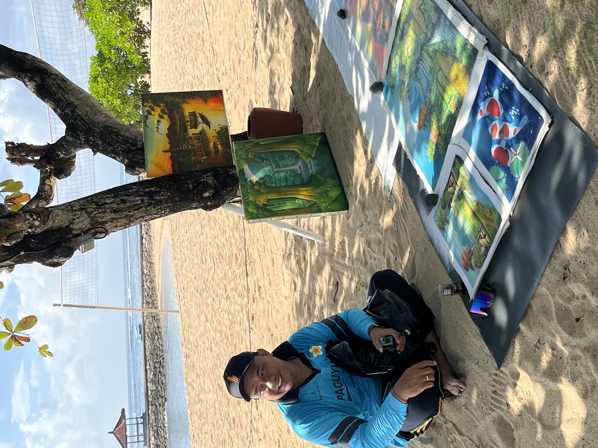 The image depicts an outdoor art display on a sandy beach, likely in Kuta Selatan, Indonesia, with the ocean and a gazebo visible in the background. A man wearing a blue polo shirt and a black baseball cap is seated on the sand, holding a black object, possibly a camera. In front of him are several paintings laid out on a black mat, some propped up on easels, featuring various scenes like a sunset landscape, a waterfall in a forest, and koi fish. The environment suggests a tropical setting with palm trees and lush greenery.