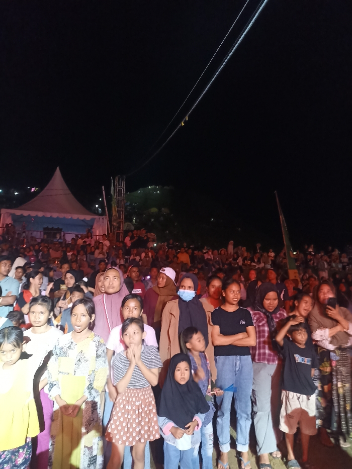 This nighttime photograph captures a large outdoor gathering in Kecamatan Pujut, Indonesia, filled with numerous people observing an event. In the foreground, a diverse group of individuals, primarily women and children, stands attentively. Many are looking forward, although some are gazing directly at the camera. Several women are wearing hijabs or other head coverings, and one woman in the mid-ground wears a light blue face mask.
The background reveals a dense crowd stretching into the distance. To the left, a large, illuminated white marquee tent with blue trim is prominently visible, suggesting a stage or central activity area. Adjacent to the tent are metal scaffolding structures. The entire scene is set under a dark night sky, illuminated by artificial light sources that highlight the attendees and the tent. Distant lights are discernible on a hillside in the far background, hinting at nearby settlements or further activity. Two distinct white cables or wires extend diagonally across the upper portion of the dark sky. The overall atmosphere suggests a community event, possibly a performance or public viewing, with attendees calmly engrossed in the proceedings. There is no clearly readable text visible on any objects or clothing.