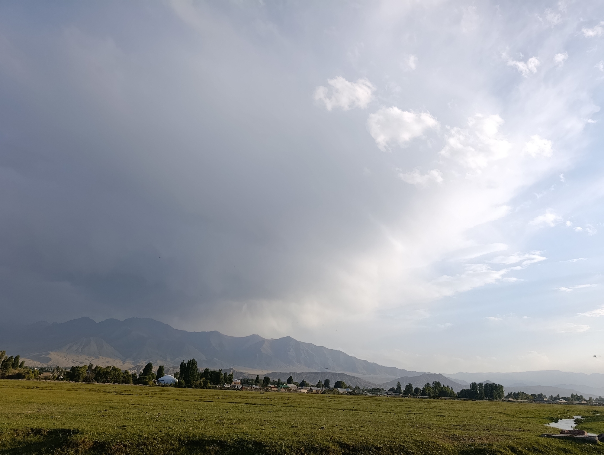 In the city of Kochkor, Kyrgyzstan, a vast expanse of green grass stretches towards a distant mountain range. The sky above is a dramatic display of contrasting weather. To the left, heavy, dark clouds loom, suggesting an impending storm, while to the right, the sky is bright and clear, dotted with scattered white clouds. A small village is nestled at the base of the mountains, with a cluster of houses and trees visible. A peculiar white dome-shaped structure stands out among the buildings, possibly a yurt. The overall scene captures a moment of transition, with the threat of rain on one side and clear skies on the other.