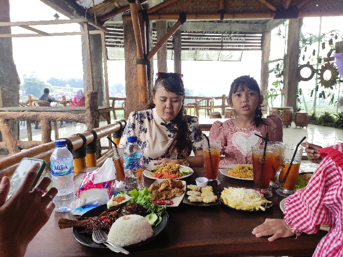 The scene is a restaurant located in Kecamatan Pujon, Indonesia, with coordinates -7.855 latitude and 112.454 longitude. There are four people seated at a wooden table. The table is covered in a wooden tablecloth. There are many dishes with food, drinks and glasses, including fried noodles, fried fish, rice, fried chicken, fries, and iced tea. Two of the people are female and have long black hair. One is wearing a floral patterned shirt and the other is wearing a red and white striped shirt with a white heart design. One of the females is looking at the camera. The third person is a woman wearing a red and white plaid shirt. She is leaning toward the table and looking at the food. The fourth person is mostly out of frame but appears to be an adult male. There is also a partially full water bottle with a blue label on the table. There are bamboo slats behind the table and to the left and right sides of the frame, and a wooden structure with bamboo panels above. The restaurant appears to have a rustic and open-air design. The weather is overcast and there is a lot of green foliage outside the window.