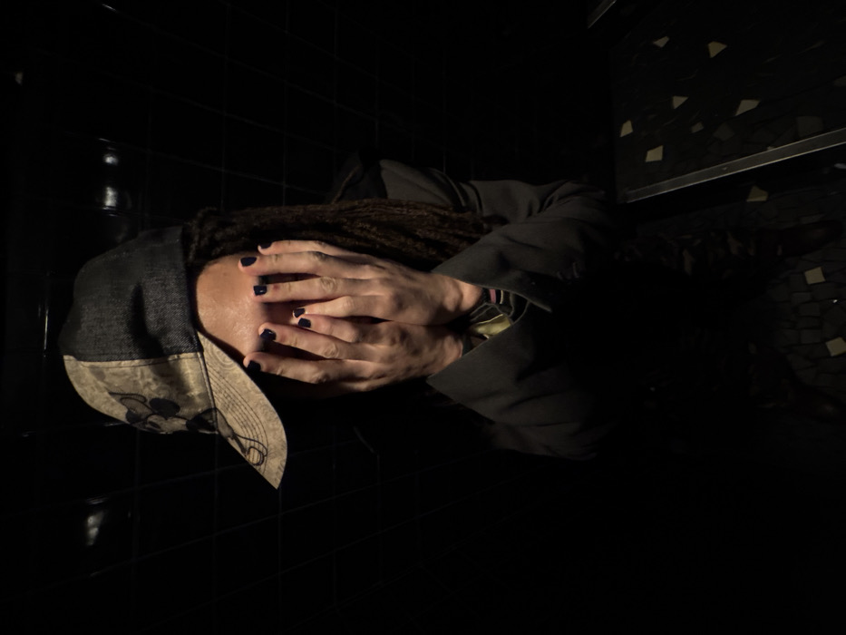 A person lies on a black-tiled surface, face obscured by hands. Dark nail polish adorns the fingers. A baseball cap with a light-colored brim rests beside the head, and dreadlocks frame the face. The individual is dressed in a black blazer or coat. The lighting is dramatic, casting much of the scene into shadow, save for where light reflects off the tile. A partially visible wall with mosaic tiles and a thin shelf can be seen in the upper right corner.