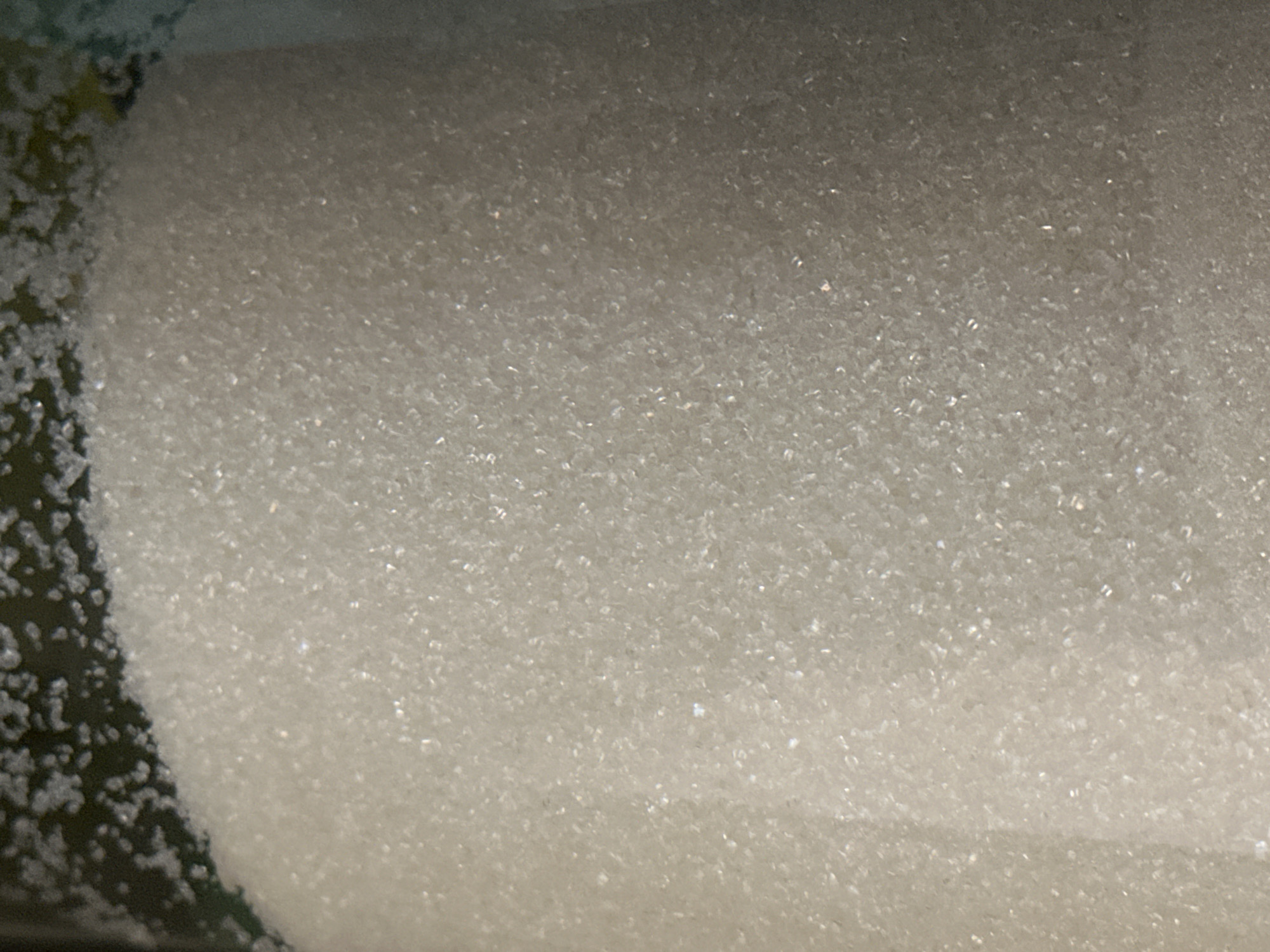 The image shows a close-up view of granulated sugar. The sugar is light in color and appears to be dry. The texture is granular, with individual sugar crystals visible. There is a dark area at the top of the image, which could be the inside of a container. There is no indication of the city of Nevinnomyssk, Russia, or any other specific location. The image is a still photograph and does not depict any actions or interactions.
