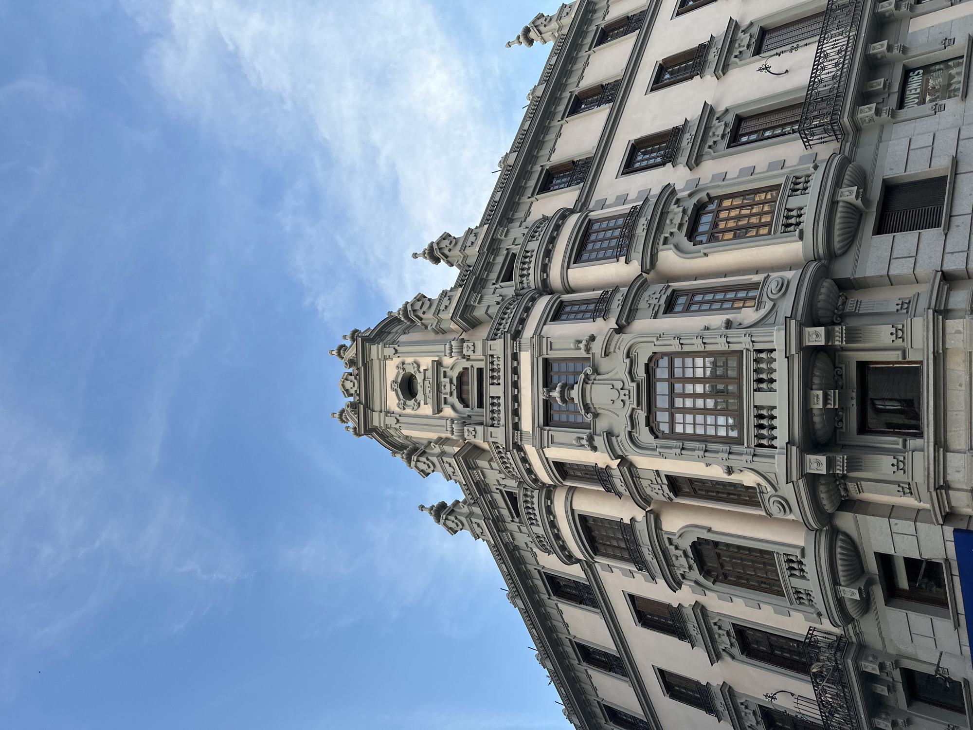 A low-angle shot shows the ornate facade of a building in Madrid, Spain, set against a blue sky with wispy clouds. The building is tall and features multiple stories with windows framed by intricate stonework. Architectural details include balconies with metal railings, decorative moldings, and embellishments around the windows. A tower-like structure is at the building's corner, with a circular window and additional decorative elements at the top. The building's exterior is light in color, possibly off-white or pale gray, with contrasting darker details. The angle of the shot gives a sense of the building's height and grandeur. A small sign is visible near the building's edge.