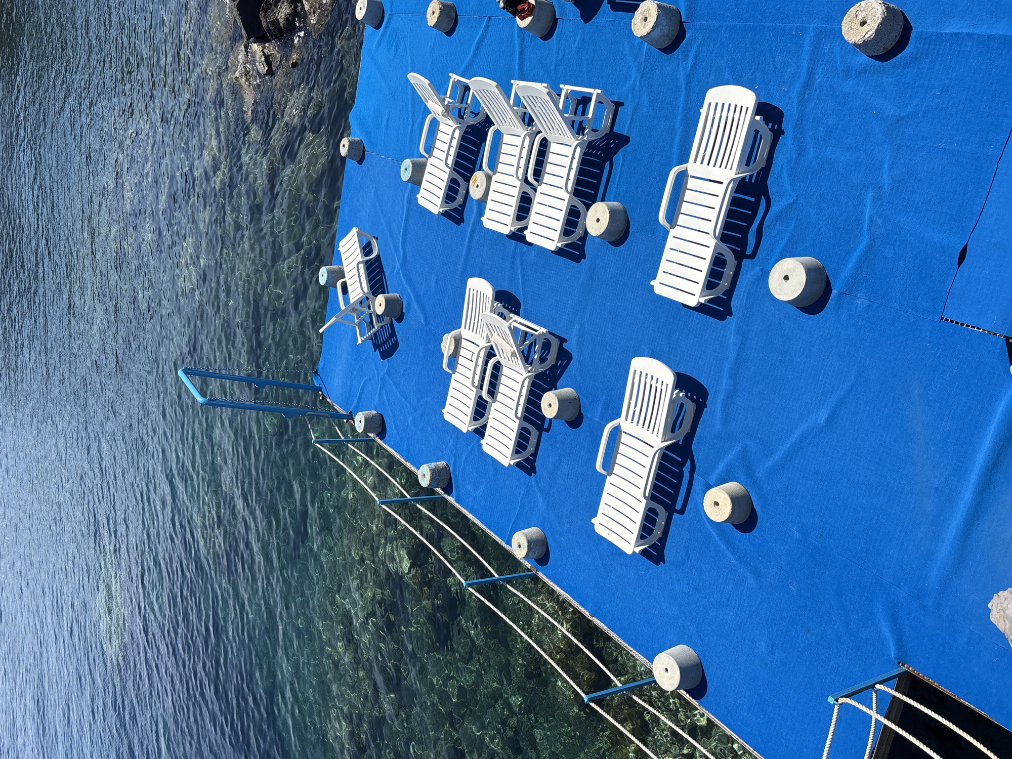This is an overhead shot of a bright blue floating platform alongside clear, shallow coastal water. The platform is furnished with numerous white plastic lounge chairs or sunbeds, some partially folded, others fully extended, all casting distinct, sharp shadows that indicate strong overhead sunlight, likely midday or early afternoon.
Several grey, cylindrical concrete weights are positioned across the blue platform, some with ropes attached, likely serving to anchor or stabilize the floating structure. Along the edge of the platform facing the water, a white rope barrier is strung between small blue posts, with additional concrete weights intermittently spaced. A blue ladder-like structure extends into the water from the platform.
The water next to the platform is exceptionally clear, revealing a rocky seabed and submerged rock formations. Ripples are visible on the water's surface, reflecting the sunlight. The water transitions to a deeper, darker blue further away from the platform. The scene suggests a tranquil leisure area, possibly a private dock or a section of a resort or beach club, emphasizing relaxation by the sea. No individuals are visible, and there is no visible text in the image. The setting is consistent with a sunny, coastal environment in Lipari, Italy.