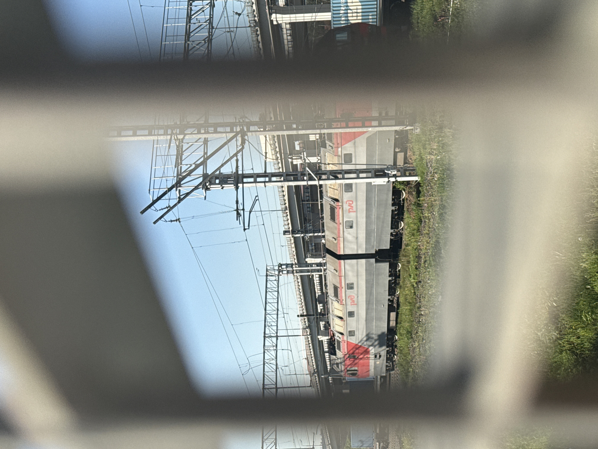 A train of gray and red passenger cars is situated on a track with overhead electrical lines. A green, grassy area extends alongside the track. A portion of a light-colored structure obscures the view of the scene. The word "PZD" is visible on the train car. The location is Moscow, Russia.
