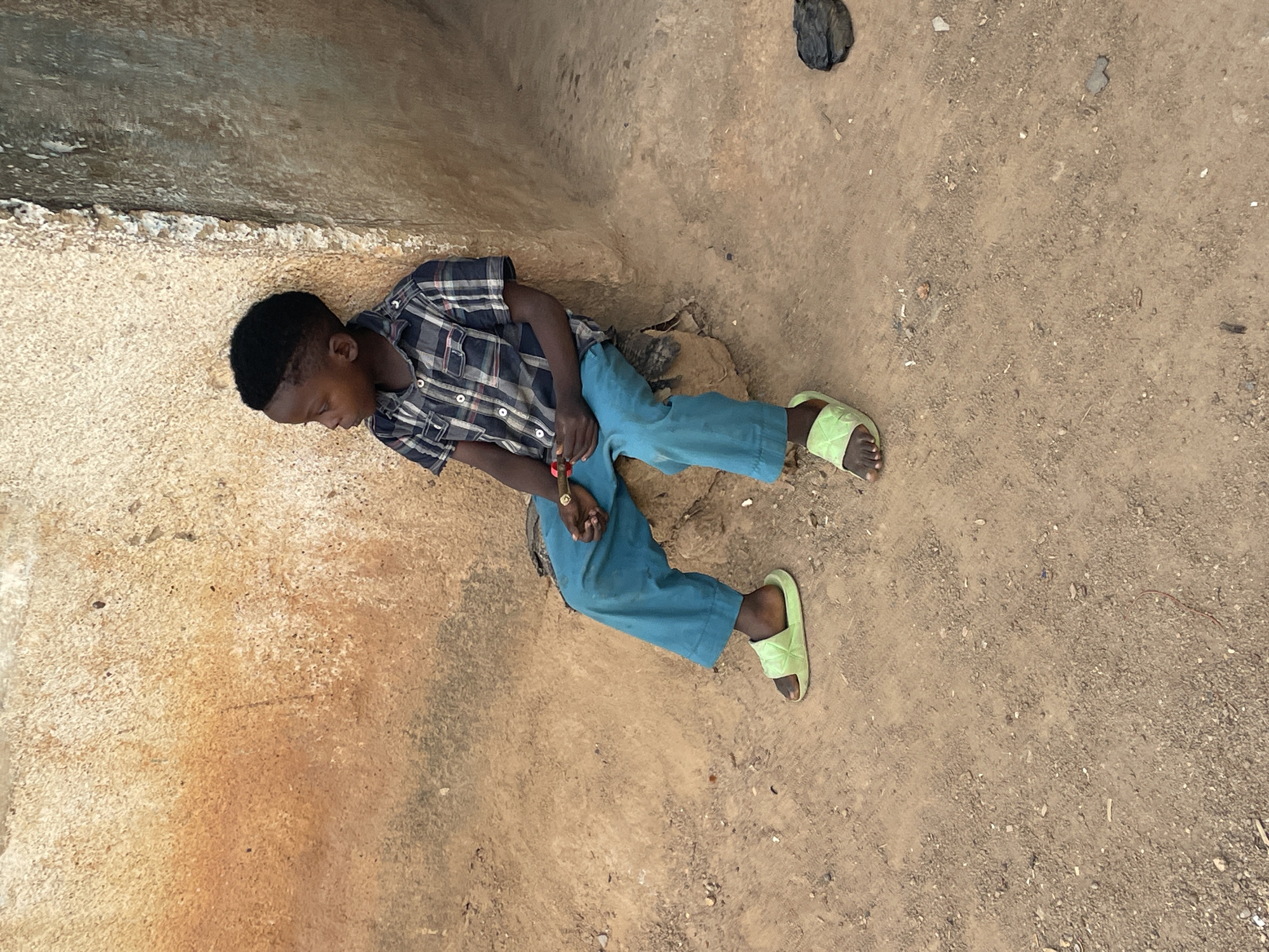 A young Black boy wearing a checkered short-sleeve shirt, blue trousers, and light green sandals is lying on the ground in Jos, Nigeria. He is positioned in the corner where two light-colored, textured walls meet. A black plastic bag sits on the sandy ground near the top right corner of the frame.