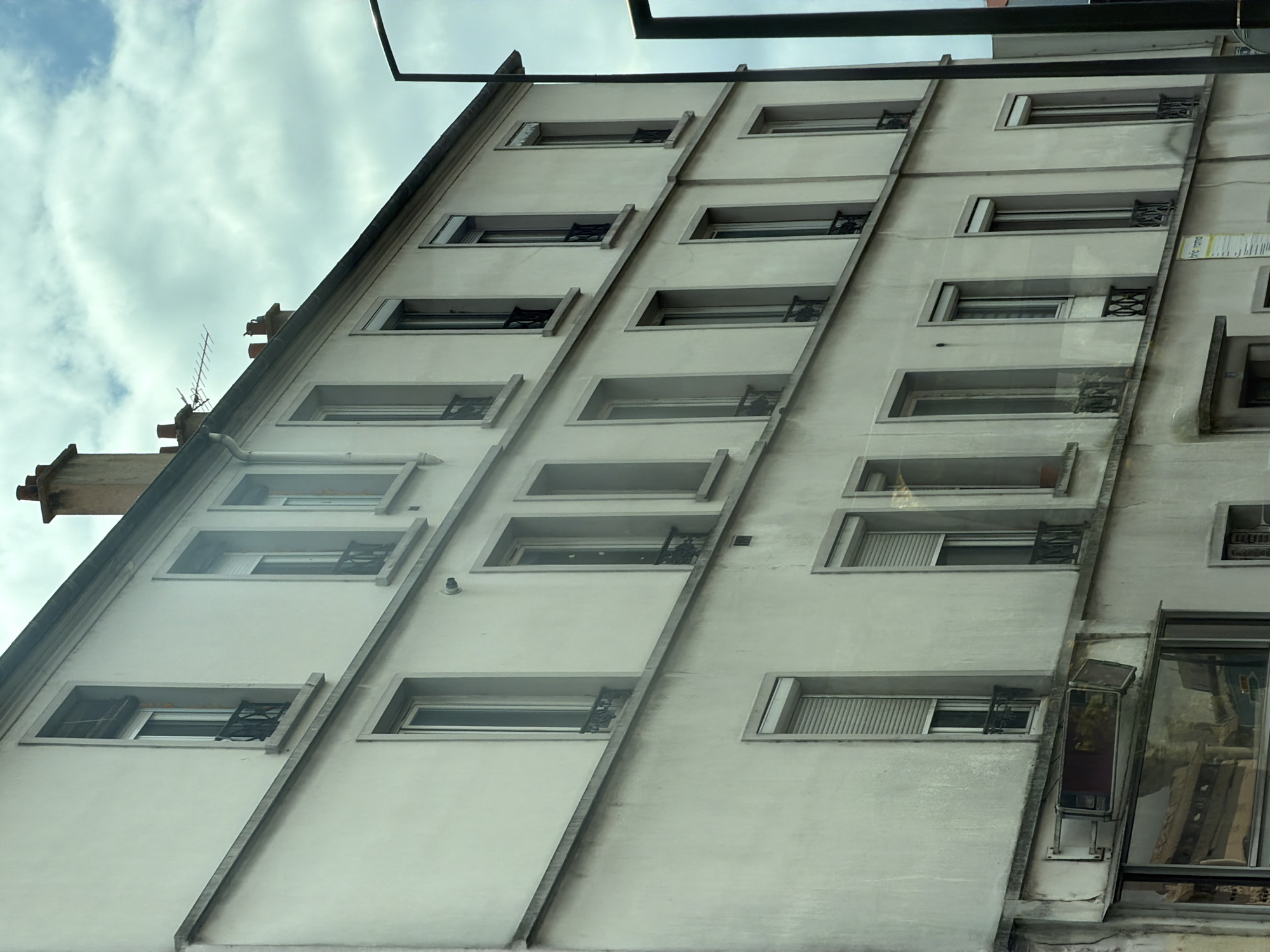 The image shows the side of a multi-story building with a cream-colored facade, angled towards the viewer. The building features numerous rectangular windows, many with dark metal security bars. The windows are arranged in a repeating pattern across the facade, with some appearing to be open. There are also external architectural elements like ledges and a diagonal concrete beam running down the side.
In the upper left of the image, a section of a darker, sloped roof is visible, along with a chimney stack and what appears to be an old television antenna. The sky above is cloudy with patches of blue. Reflections are visible in some of the windows, suggesting the photo might have been taken from across a street or an opposing building. The lighting suggests it could be daytime, possibly overcast.
On the lower right, a larger window displays a reflection of buildings across the way. There is also a small, metallic rectangular object mounted on the wall below this window. A vertical strip of text, appearing to be a label or sign, is visible on the right side of the building, between rows of windows. The overall scene depicts an urban residential or commercial building exterior in Alfortville, France. No people are visible in the image.