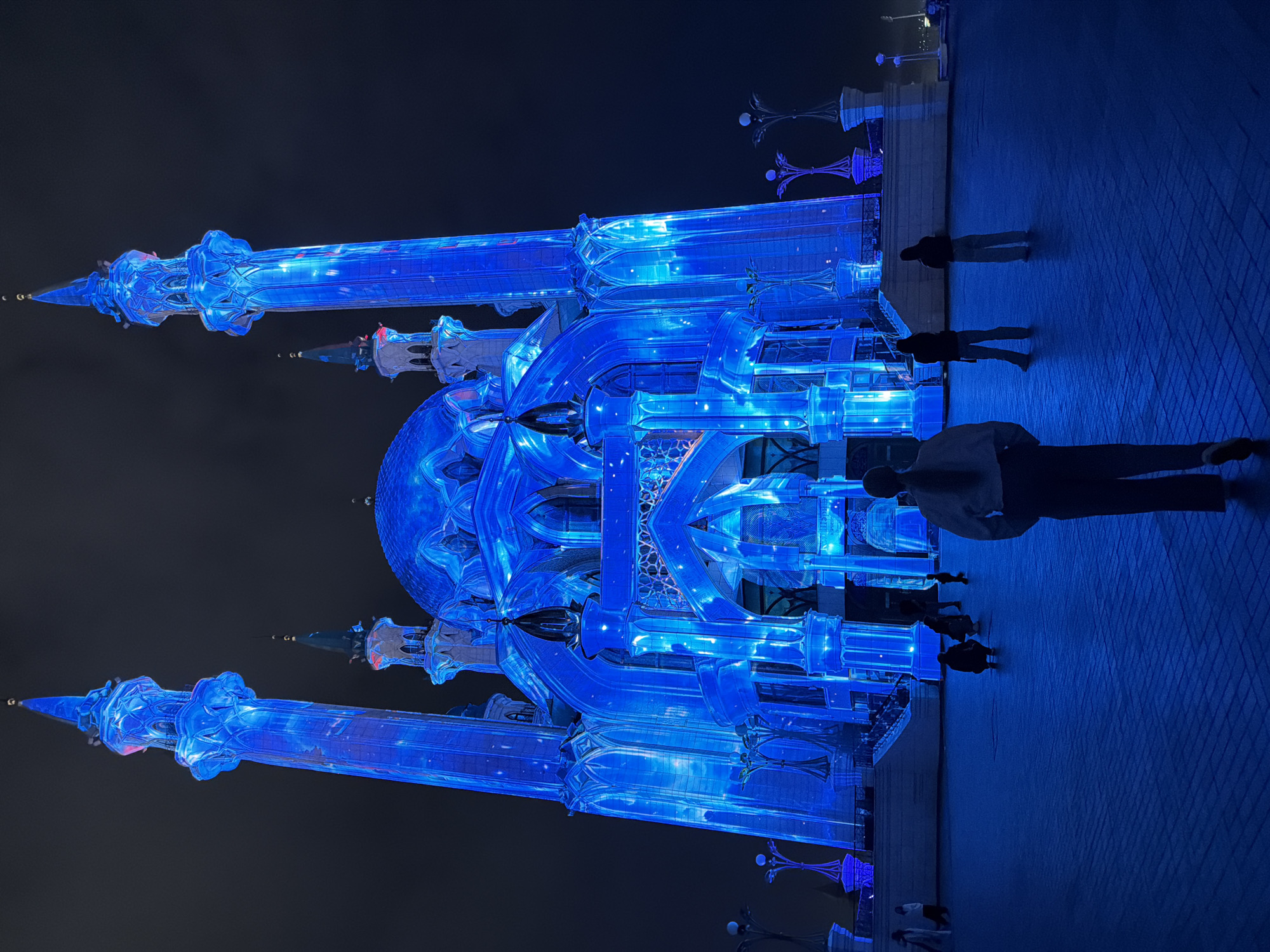 Captured at night in an unknown city, this image presents a blue-lit architectural structure, likely a mosque, positioned next to a tiled plaza where several pedestrians are present. The structure features multiple turrets and a central dome illuminated in blue, creating a striking contrast against the dark sky. A group of people are seen standing and walking on the plaza, which is illuminated by streetlights along the periphery.