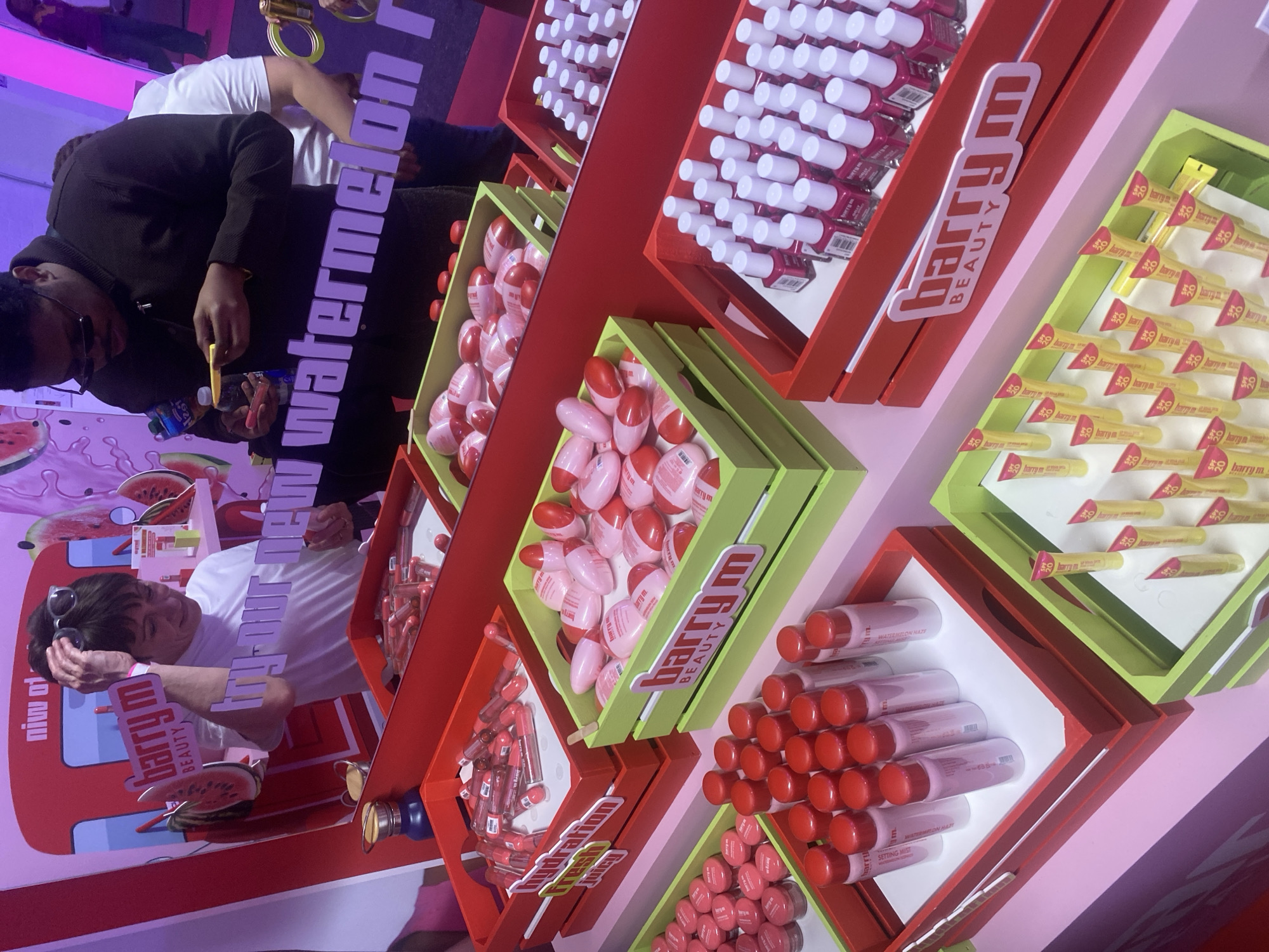 This dynamic, tilted image captures an engaging beauty product display, likely at a promotional event or pop-up store in London, United Kingdom. The scene is dominated by vibrant red and lime green display crates filled with various cosmetic items from the brand "Barry M Beauty."
Several types of products are showcased: rows of nail polishes with white caps in red tiered boxes, numerous pink and red egg-shaped lip balms or tints, and yellow tubes of "Barry M SPF 30" sunscreen in lime green trays. Another section features bottles labeled "WATERMELON HAZE SETTING MIST," hinting at a specific product line. Watermelon graphics are visible on the background, reinforcing the "new watermelon" theme, partially seen in large purple lettering.
Two men are visible, interacting with the display. On the left, a man wearing a dark jacket and sunglasses is holding a small item, seemingly examining a product. On the right, another man in a white t-shirt, with sunglasses perched on his head, looks towards the left side of the frame. The overall atmosphere is lively and colorful, enhanced by pink and purple lighting that bathes the indoor setting. The prominent branding "Barry M Beauty" is seen on multiple display elements, alongside keywords like "hydration" and "fresh." The image, taken from a high angle, creates an immersive view of the promotional setup.