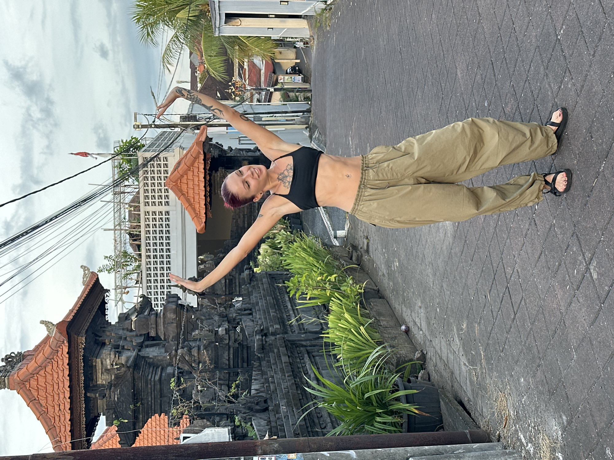 A full-body shot of a woman with pink-tinted short hair, wearing a black sports bra and khaki pants, is oriented sideways on a paved street in Dalung, Indonesia. She stands with arms outstretched. A traditional Balinese building with a dark-stone facade and a tile roof is to her left. To her right, across the street, is a building with a light exterior and an adjacent palm tree. Overhead power lines are visible. The street is gray and tiled. Vegetation grows along the building on the left side of the street.