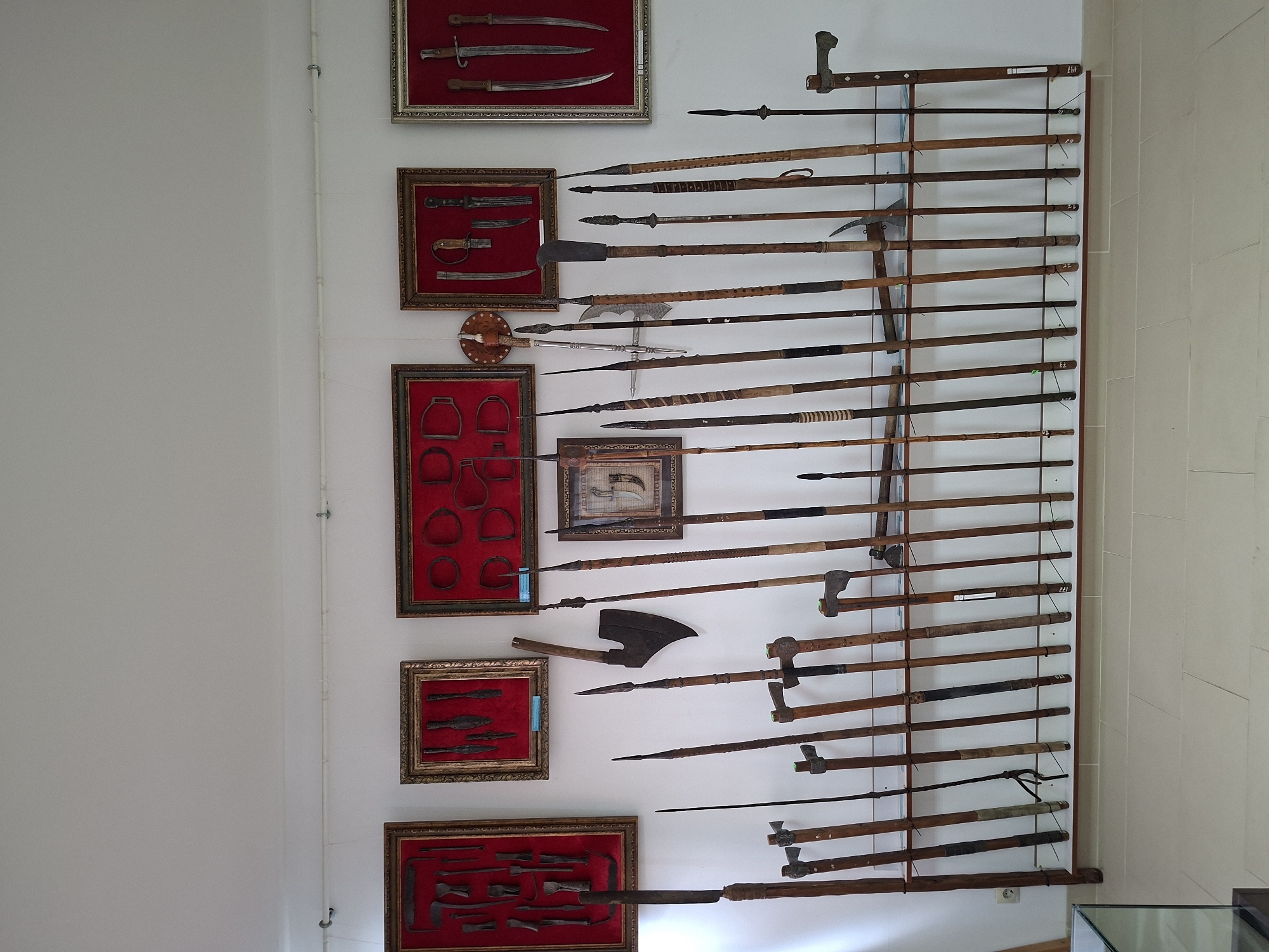 The image depicts a museum exhibit displaying a collection of historical weapons, likely in Ialoveni, Moldova. The items are arranged on white walls and a long wooden shelf against a white background.
The exhibit features several framed displays on the walls. The leftmost framed display contains various metal tools and blades against a red background. The next framed display to the right also holds metal objects on a red background. A smaller, ornate framed piece is centrally located, showcasing what appear to be arrows and possibly a small shield. To the right of this, another framed display holds several knives and daggers against a red background. The rightmost framed display contains more blades.
Below these framed items, a large collection of polearms and axes are mounted on a long wooden shelf. These weapons, predominantly made of wood and metal, vary in size and design, with visible spearheads, axe heads, and other bladed implements. Some of the wooden shafts are wrapped with different materials, and some have markings or labels.
In the foreground, a glass display case is partially visible on the left side, suggesting a more extensive museum setting. The flooring appears to be tiled. The overall environment is well-lit and organized, characteristic of a museum exhibition. No people are visible in the image.