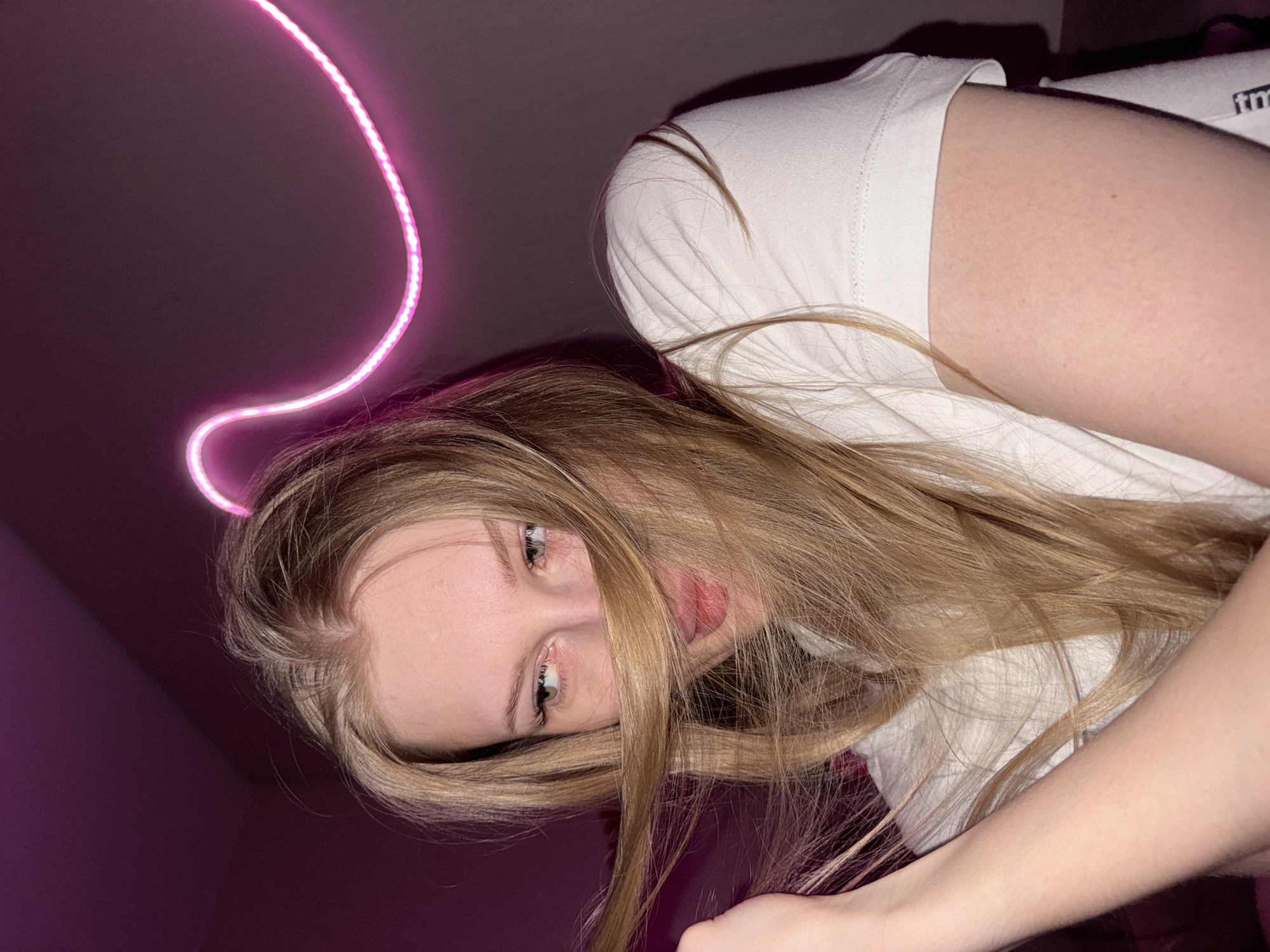 This is a close-up, eye-level, medium shot of a young Caucasian woman with long blonde hair and light-colored eyes. She is wearing a white t-shirt. Her hair is partially obscuring her face, and she is looking to her left. Her left arm is visible, holding some of her hair. In the background, a neon pink light fixture is visible on a dark wall, which is illuminated with purple light. The image was taken indoors, likely in a residential setting, and does not contain any elements indicating a specific location in the city of Perm, Russia.