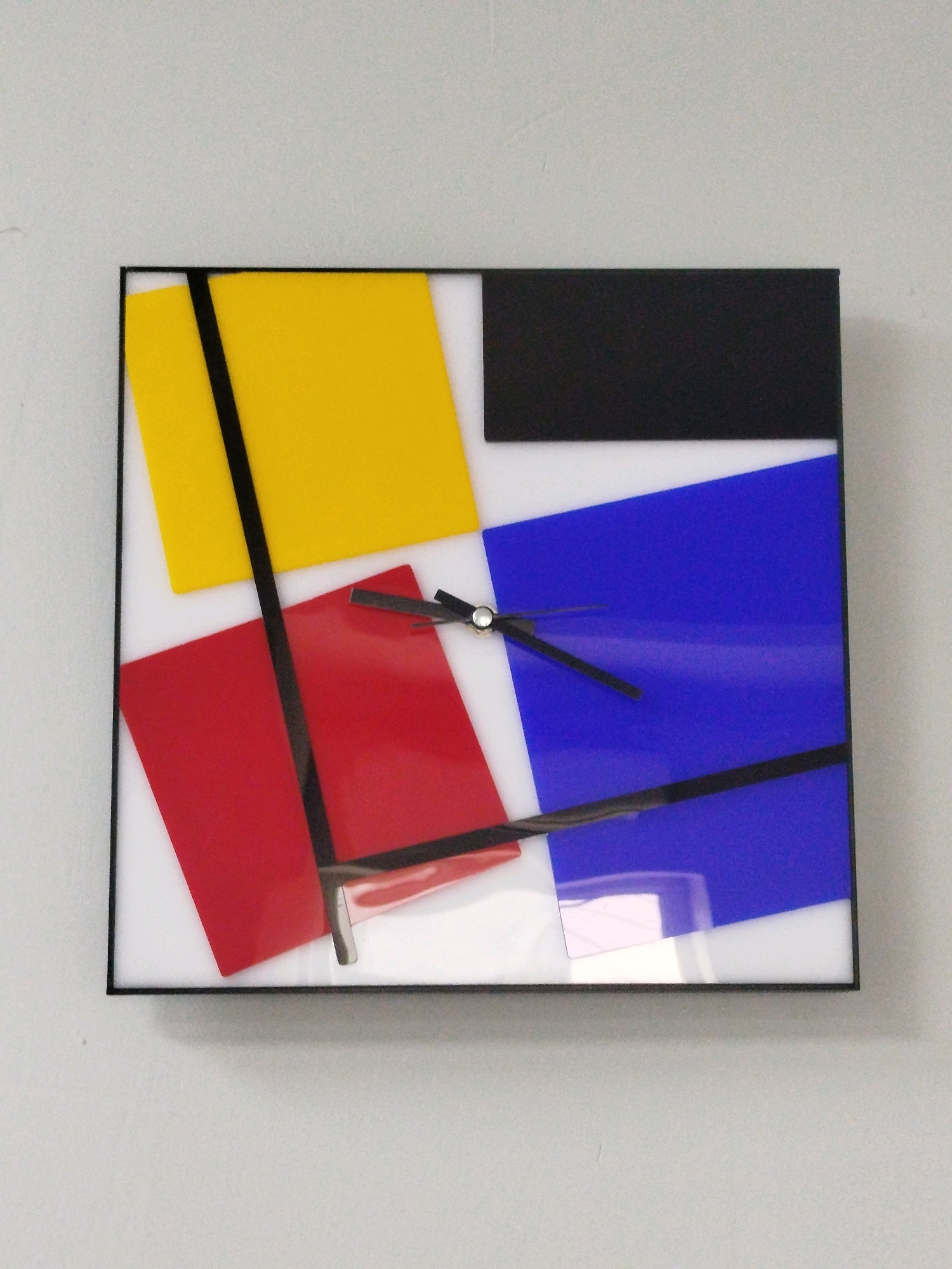 A square clock with a black frame hangs on a pale gray wall. The clock face is abstract, featuring colored rectangles and black lines. A yellow rectangle is in the upper left, a black rectangle is in the upper right, a red rectangle is in the lower left, and a blue rectangle is in the lower right. Black lines divide some of the colored rectangles. The clock hands, black and thin, are visible near the center of the clock face, pointing to approximately 10:07. The overall style is reminiscent of Piet Mondrian's neoplasticism. The clock is the only object clearly visible in the image. The lighting is even, suggesting ambient indoor lighting. There are no people or other environmental details visible beyond the clock and the wall it hangs on. The location of this clock in Tashkent, Uzbekistan is not discernible from the image.