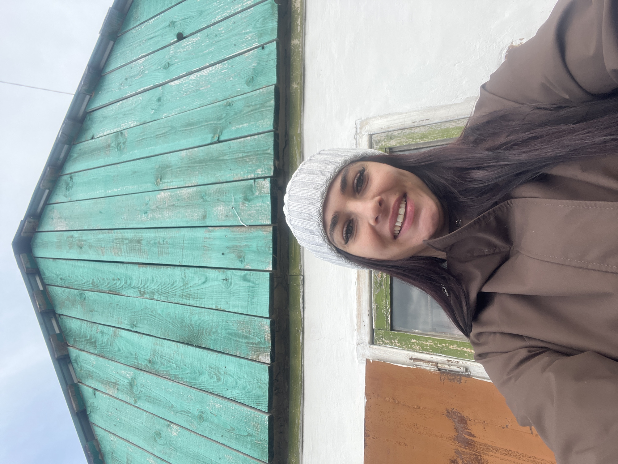 A woman is posing for a photo, with a building serving as the backdrop. She is wearing a light grey knitted hat and a brown coat. She is smiling and has long, dark hair.
The building behind her has a distinctive color scheme. The section to the left is painted in a vibrant turquoise color, while another part is white. There is a window with a green frame. Below the window is a section painted in brown. The building appears to be in a rural setting.
Given the prompt's request, it is speculated that the photo was taken in Chaikovsky, Russia.