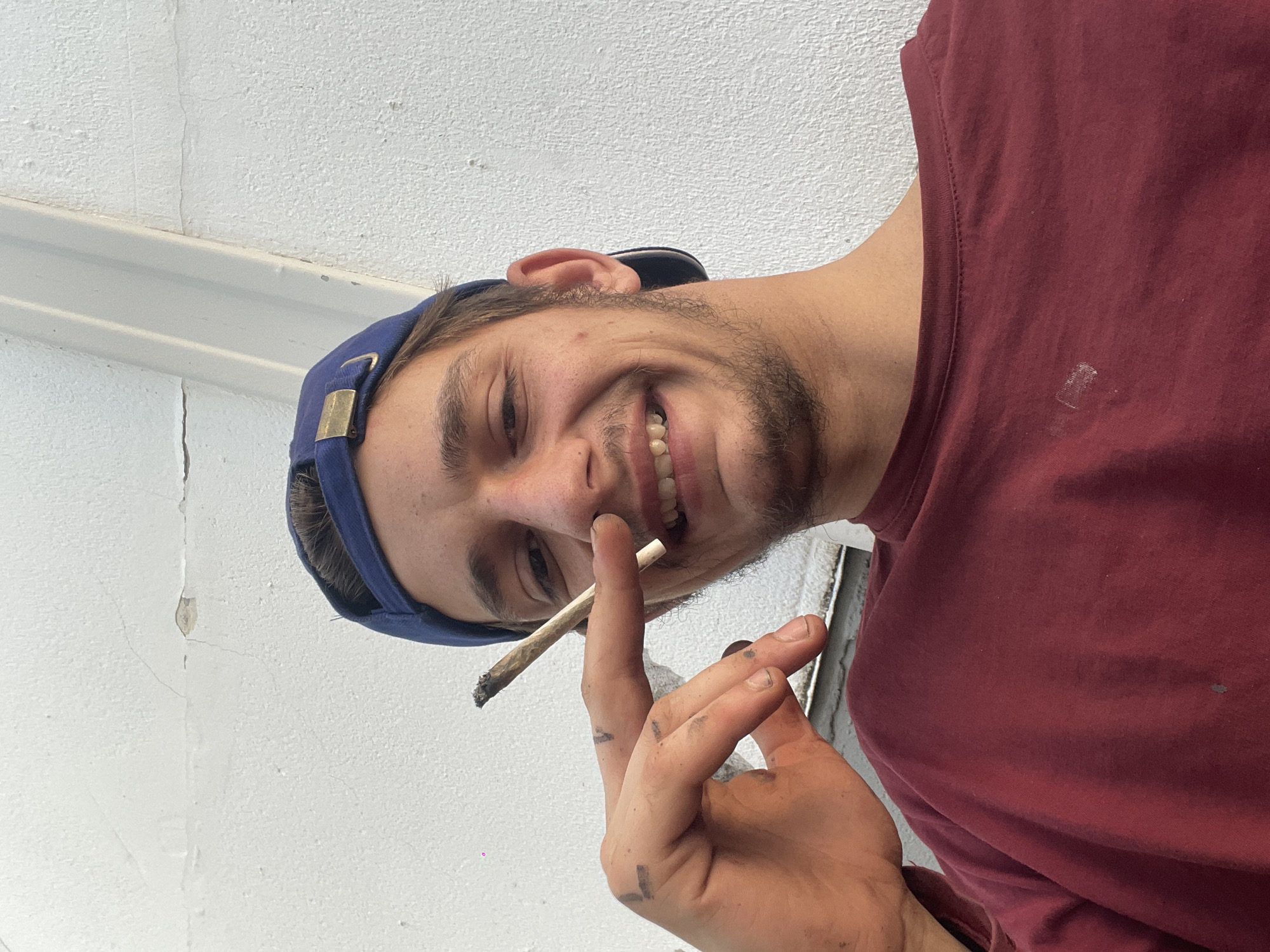 A person is shown from the chest up. The person has fair skin, a slight beard, and dark hair, and is wearing a blue baseball cap. He is smiling with visible teeth. He is also wearing a dark red t-shirt. He is holding a lit cigarette or a joint between his fingers. His hands are dirty. The background appears to be a white wall. There are no indications of any location or city.