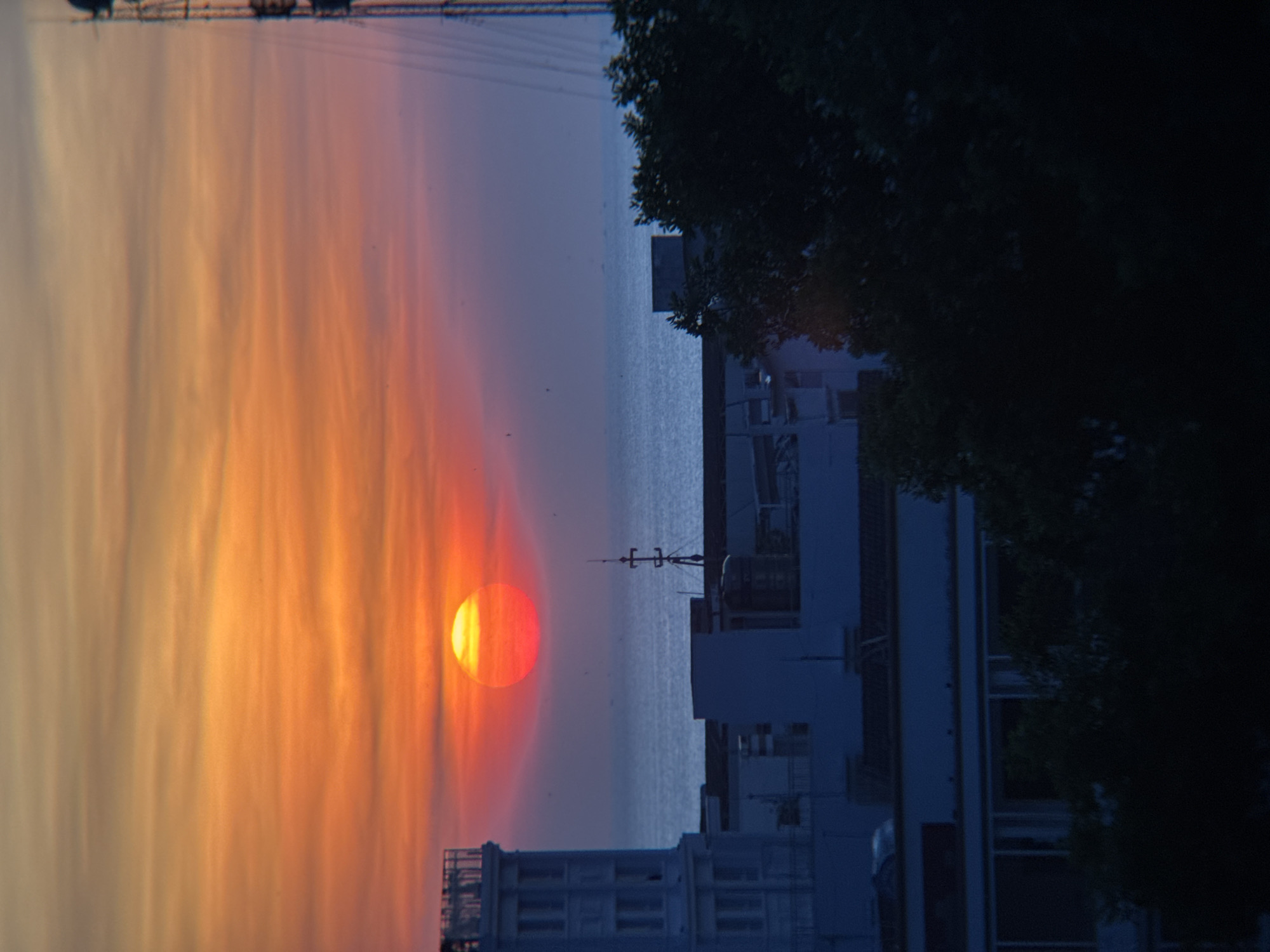 The image captures a sunset over the city of Nha Trang, Vietnam. The sun, a vibrant orb of red and yellow, is setting near the horizon, casting a warm glow across the sky. Wispy clouds streak the sky in shades of orange and purple, transitioning to a deep blue near the top. The ocean stretches out beyond the city, appearing calm and vast. In the foreground, several buildings are visible, including a white, multi-story structure on the left and darker, less defined buildings in the middle and right. Trees partially obscure some of the buildings on the right. A tall communication tower stands on the far right of the frame. The overall impression is one of a serene evening in a coastal city.