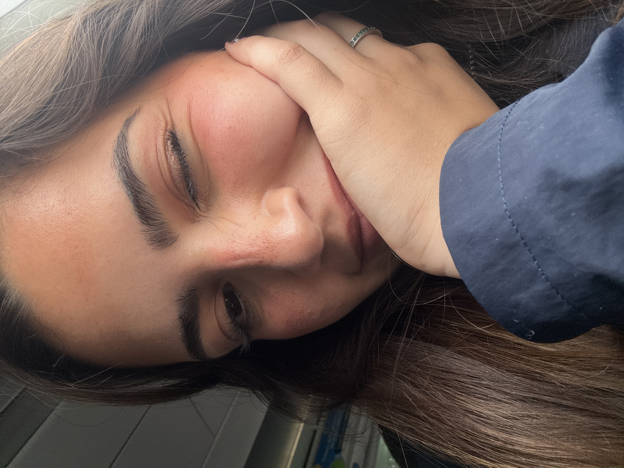 A close-up, slightly angled portrait shows a woman with brown hair and light skin. Her right hand is partially obscuring her face, with fingers pressing into her cheek. She has dark eyebrows and one eye appears closed or squinting. Her cheek has a reddish hue. She is wearing a ring on her finger. Part of a navy blue fabric, possibly clothing, is visible on the right side of the image. The background is blurred and indistinct. There is no information in the image to suggest the location of the city of San Antonio, United States of America.