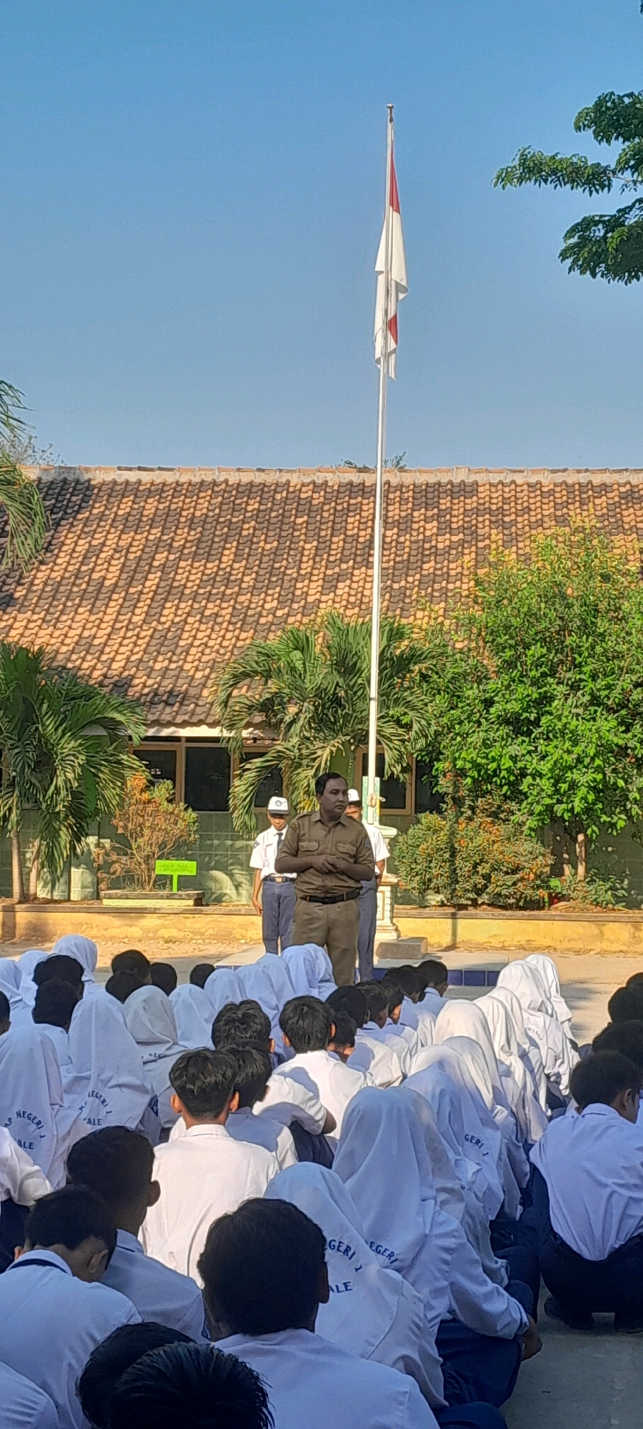 This is an outdoor, eye-level, full-shot photograph taken in daylight. A man in a light brown uniform stands in front of a group of students sitting on the ground. He is speaking and gesturing with his hands. The students are dressed in white uniforms with some wearing headscarves. Behind the man and students, a school building with a tiled roof is visible, as are palm trees and a flagpole with the Indonesian flag. The scene appears to be a morning flag ceremony at a school in Tahunan, Indonesia.