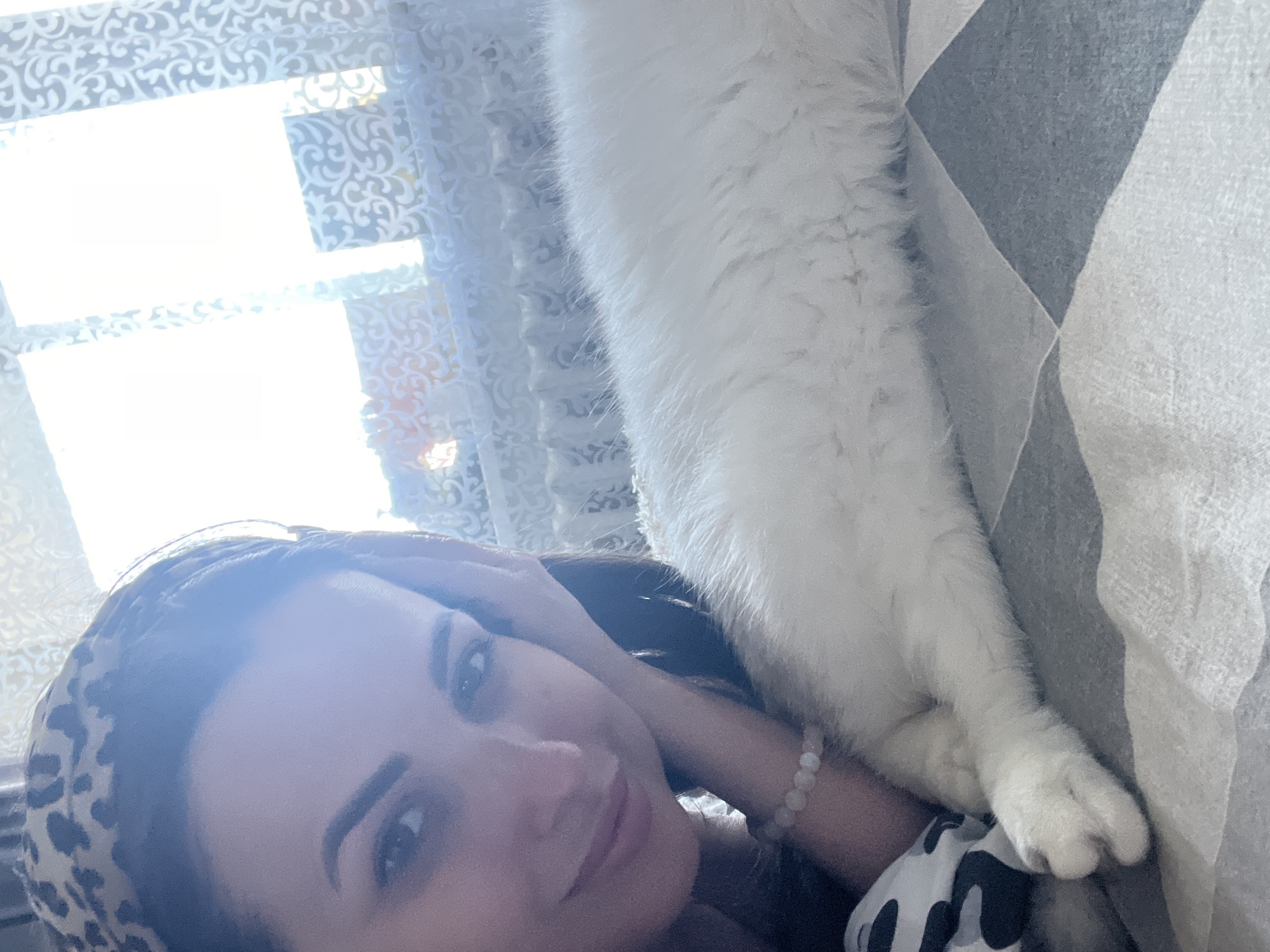 The image shows a woman lying down with her head on her hand, looking at the camera. A white cat is lying on top of her, with its long body stretching across the frame. The woman is wearing a leopard-print headband and a black and white patterned top. She has dark hair and makeup. The cat has white fur and appears to be relaxed. They are indoors, with a window covered by a patterned curtain in the background. Sunlight is streaming through the window, creating a bright, soft light. The overall mood of the image is intimate and relaxed, suggesting a moment of shared comfort between the woman and her pet. The patterned curtain and the diamond-patterned fabric the cat is lying on suggest a domestic setting. The time of day is likely daytime due to the sunlight. There is no visible text in the image.