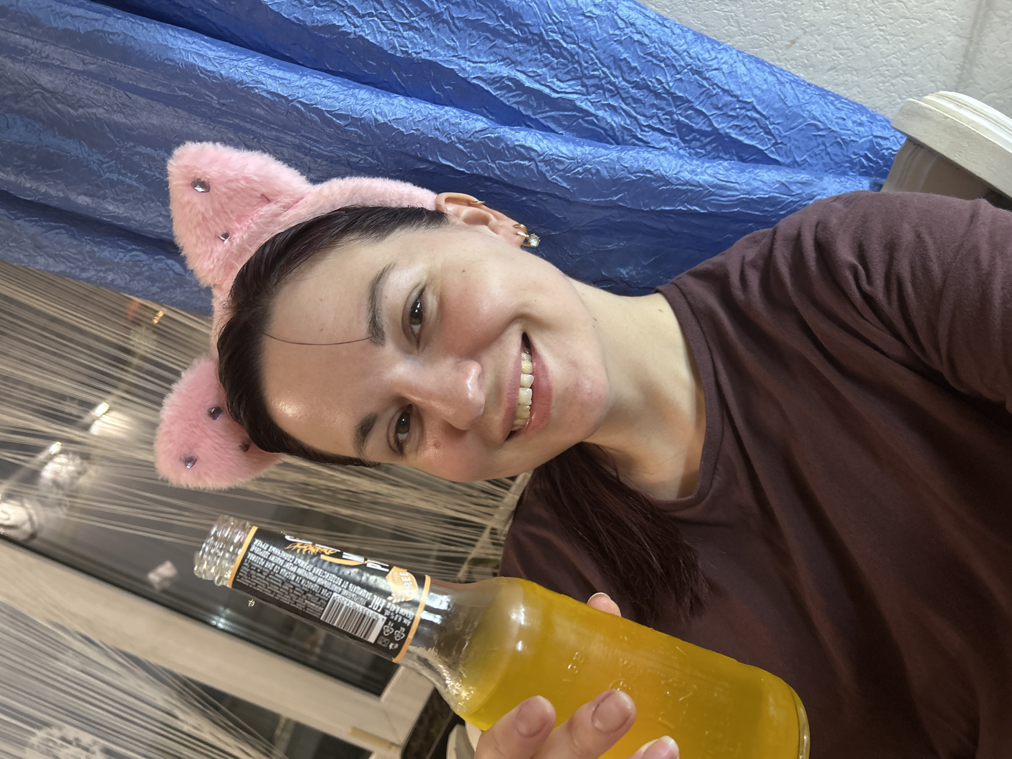 A young woman, with dark hair and a brown t-shirt, smiles and holds up a bottle of yellow liquid to the camera. She is wearing a pink furry headband with cat ears, adorned with small rhinestones. The background consists of a textured blue fabric and a window with vertical white blinds. It is not possible to determine the specific location within the city of Фоки, Russia.