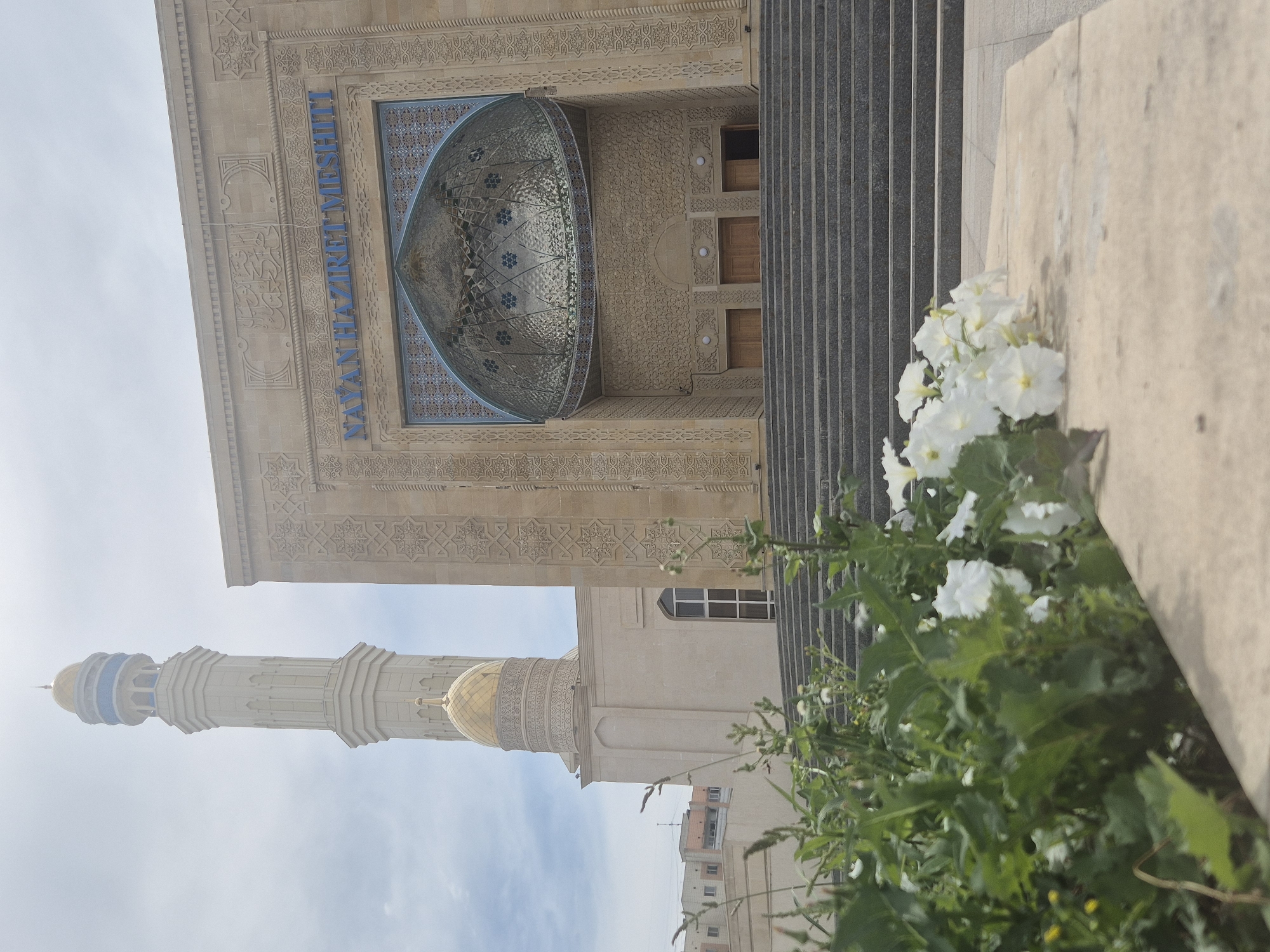 The image shows the exterior of the Nayan Hazrat Mosque in Kokshetau, Kazakhstan. The mosque features a tall, cream-colored minaret topped with a gold and blue dome, and a main building constructed of beige stone with intricate carvings. A large, ornate blue and gold dome is visible above the entrance. A wide set of stone stairs leads up to the mosque's entrance. In the foreground, a bed of green plants and white flowers is in bloom, with a stone planter box in front of them. The sky is overcast and a few buildings are visible in the background. There are no people or animals in the image.