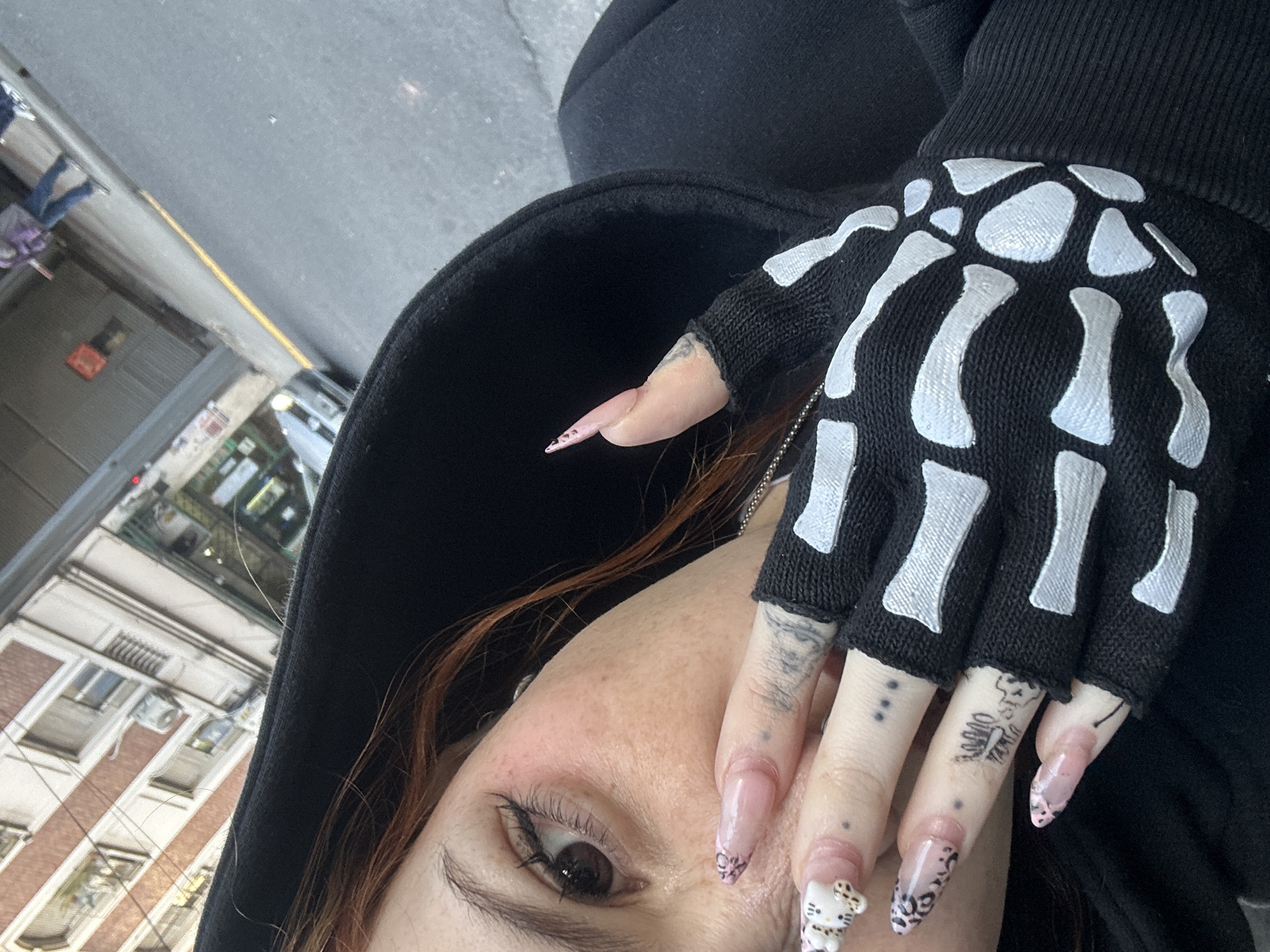 A close-up, selfie-style photograph depicts a person in a black hooded garment with part of their face and hand visible. The subject's left eye, adorned with makeup, is visible beneath auburn hair. The hand, wearing a fingerless black glove with a white skeleton bone design, is prominently displayed. The fingernails are long and adorned with various designs, including polka dots and a Hello Kitty figure. Tattoos are visible on the fingers, consisting of dots, lettering, and graphic symbols. The background shows a city street with buildings, pedestrian traffic, and cars. The architecture and street style do not definitively indicate Buenos Aires, Argentina.