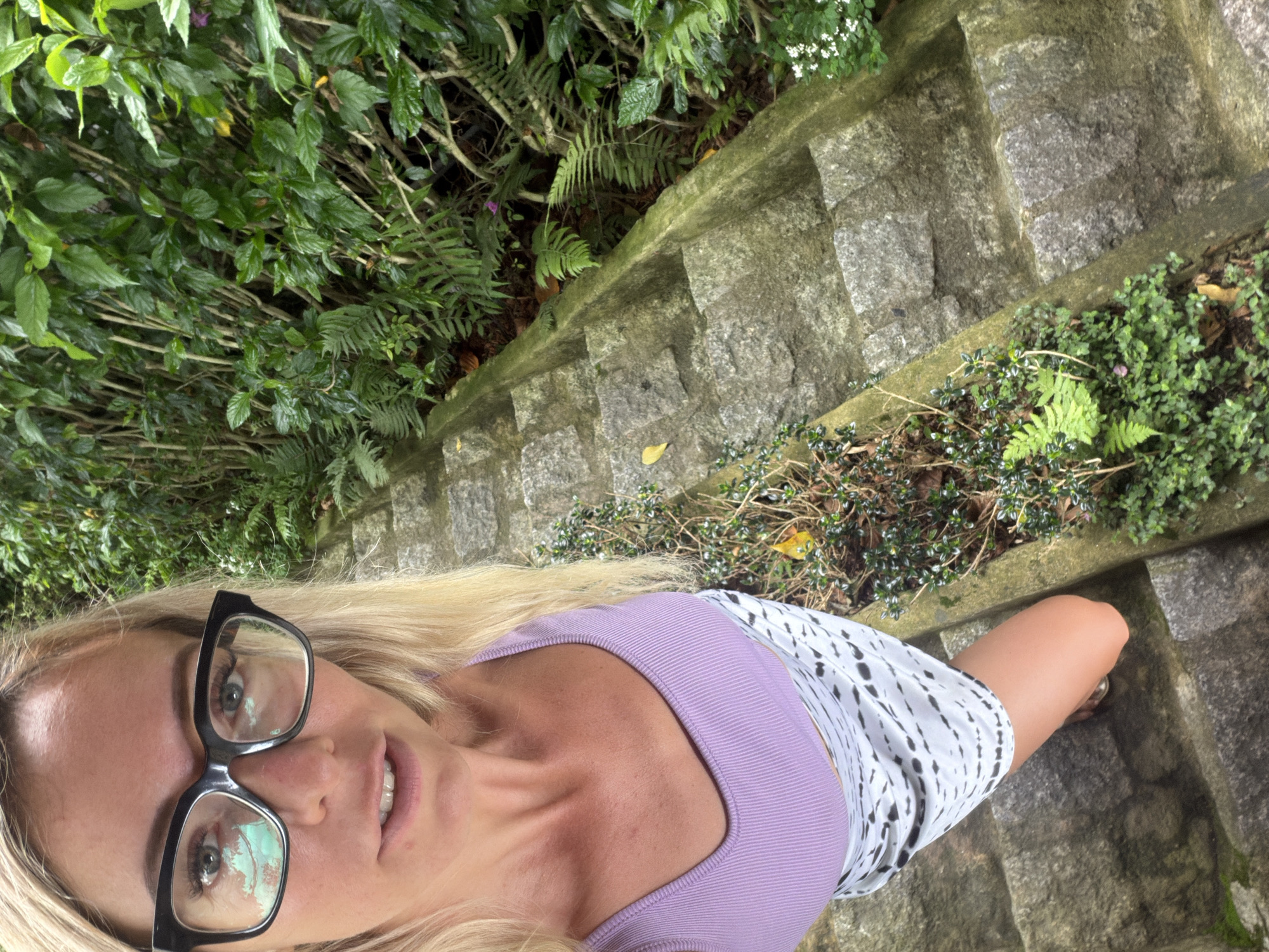 A high-angle, medium shot shows a blonde woman with dark-rimmed glasses and a purple tank top standing on a stone staircase. The woman is looking directly at the camera with her mouth slightly open. Her right hand is not visible. She is wearing a white and black patterned skirt. The staircase is flanked by lush green foliage, including ferns and small flowering plants. The overall scene suggests an outdoor setting with abundant vegetation, possibly a park or garden within the city of Xuân Thành, Vietnam.
