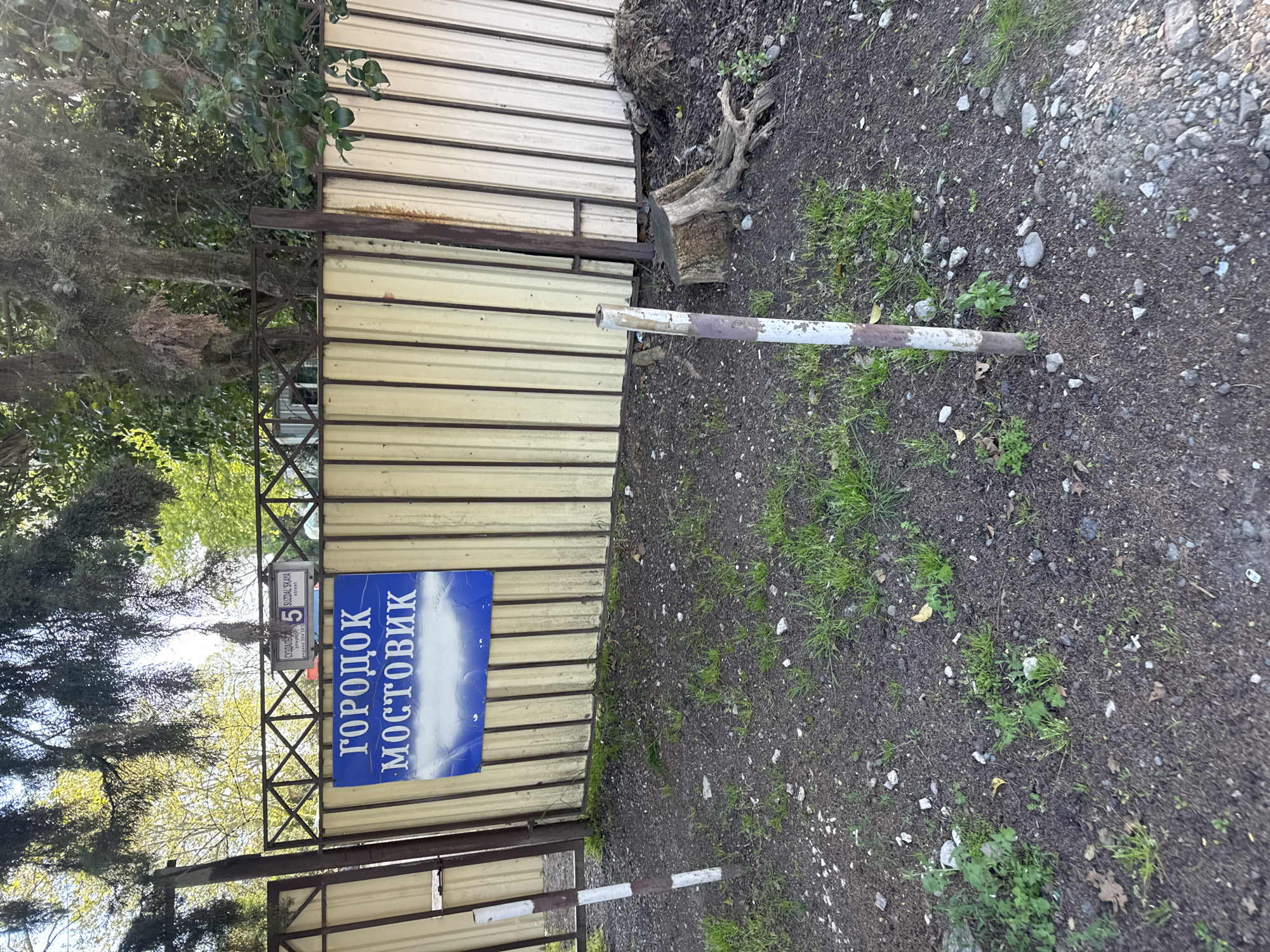 The image shows a metal gate with two blue signs written in Russian. The larger sign says "Городок Мостовик" (Gorodok Mostovik). A smaller sign above it reads "5 Suzdalskaya Street" in Russian. The gate is situated in a dirt area with patches of grass. A striped barrier is positioned in front of the gate. The background includes trees and foliage. Given the street name and the name on the blue sign, the location is likely in the city of Sochi, Russia.