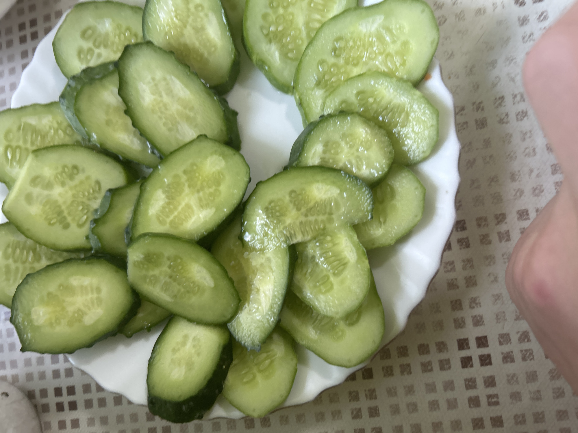 A top-down, slightly angled shot shows a white plate filled with thinly sliced cucumbers arranged in a circular pattern. The cucumbers are green with visible seeds and appear slightly glistening, suggesting they might be fresh or have been dressed. The plate is placed on a brown and beige checkered surface. A human hand, with pale skin and a pinkish tone, is visible in the lower right corner of the frame, with the thumb and index finger slightly extended, hinting at a possible interaction with the plate or its contents. The overall lighting is natural and diffused, with no harsh shadows. The scene is devoid of any explicit location markers that would identify it as being in the city of Старотумбагушево, Russia.