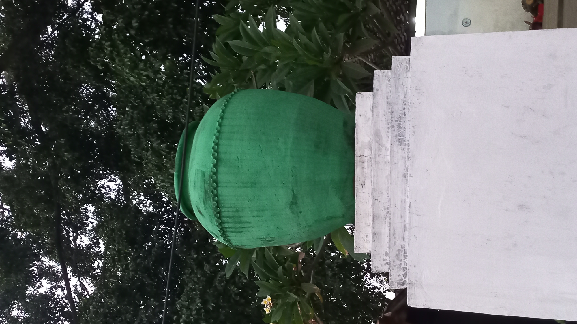 A vibrant green, large ceramic pot sits atop a multi-tiered white pedestal. The pot has a rounded body with a distinct rim and is decorated with a circular, beaded pattern just below the rim. The pedestal, made of concrete, has several horizontal layers, each appearing slightly weathered. Behind the pot and pedestal, lush green foliage from trees and other plants fills the background, with some branches extending towards the sky. A hint of a building with a tiled roof is visible in the far left background, and a glimpse of another structure with a circular emblem on its wall can be seen on the right. The scene appears to be outdoors, possibly in a garden or public space in Pare, Indonesia, given the tropical foliage and the style of the pot. No people or animals are visible in the image.